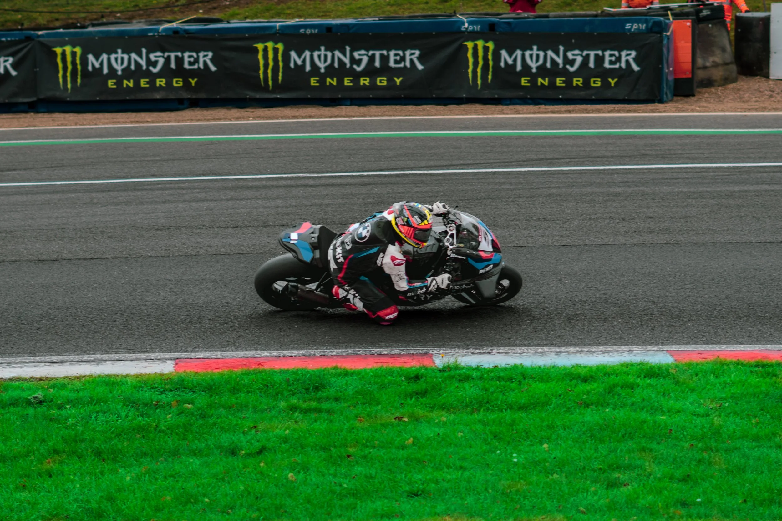 A motorcycle racer in a black racing suit riding a motorcycle on a race track with 'Monster Energy' branded barriers in the background.