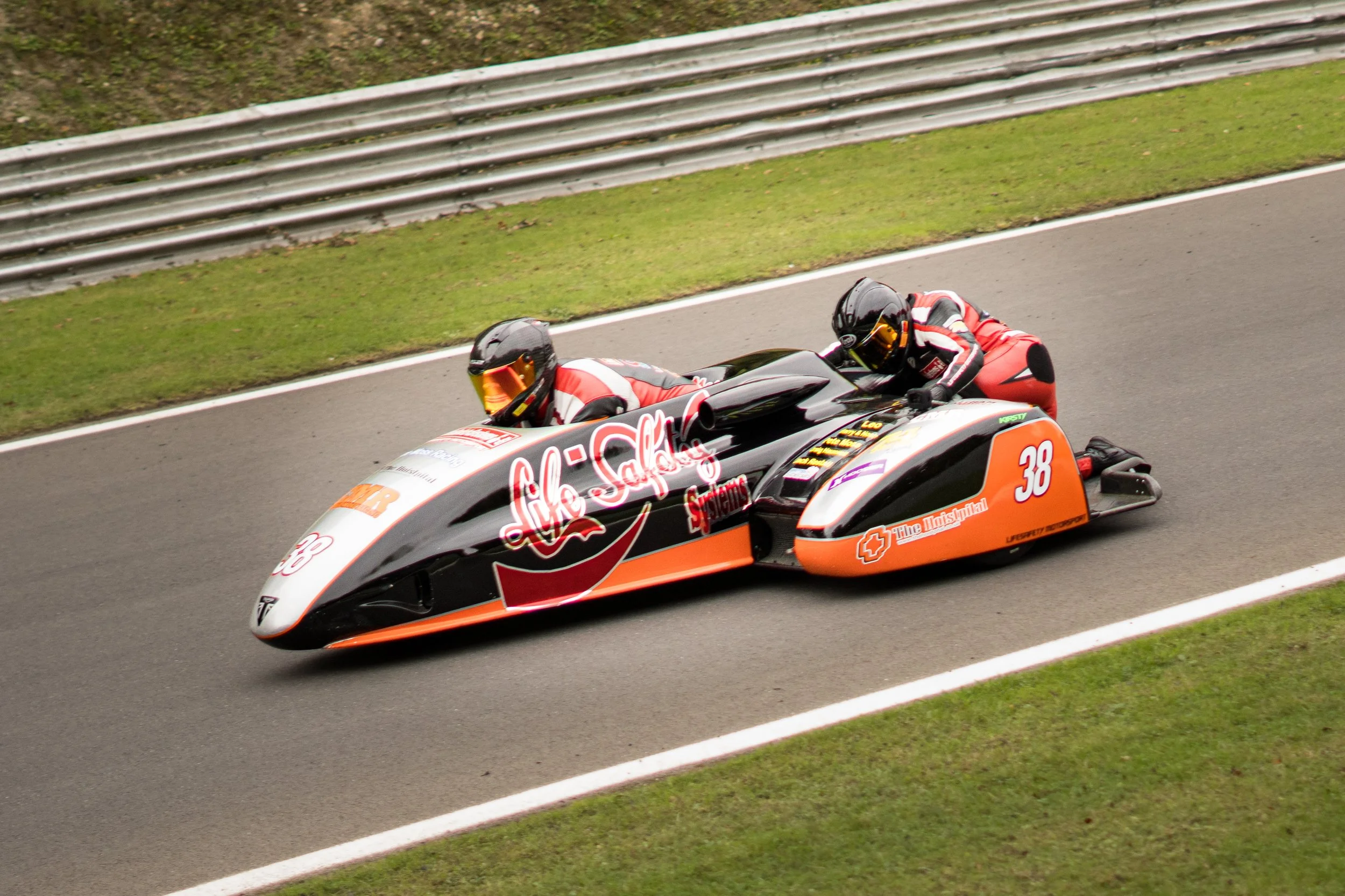 Two racers in a sidecar motorcycle racing on a track, wearing helmets and racing suits, with a grassy area and metal barriers in the background.