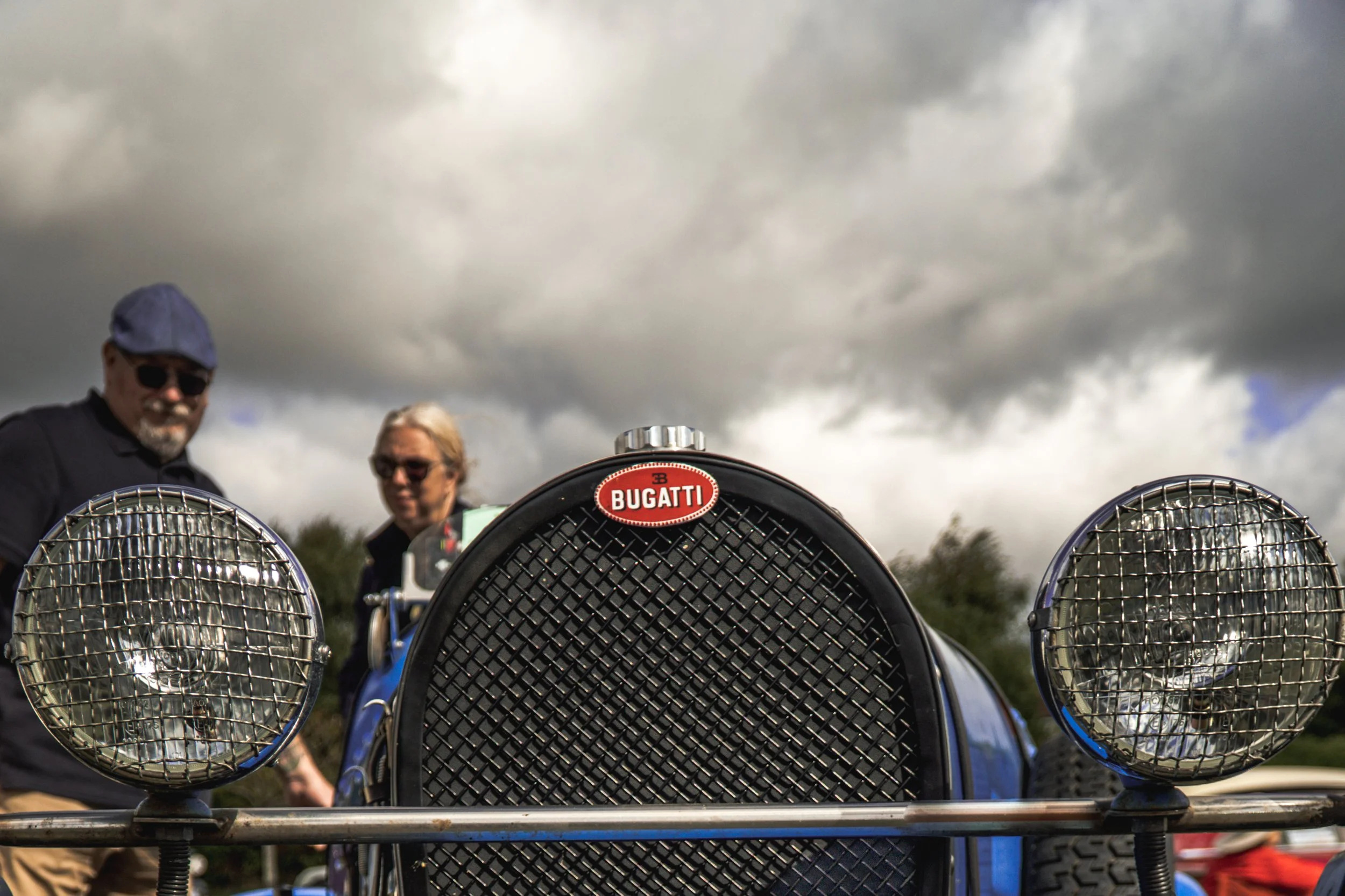 Close-up of a vintage blue Bugatti race car from the front, with three large circular headlights with metal grilles and a prominent Bugatti emblem, under a cloudy sky. Two people are visible in the background.