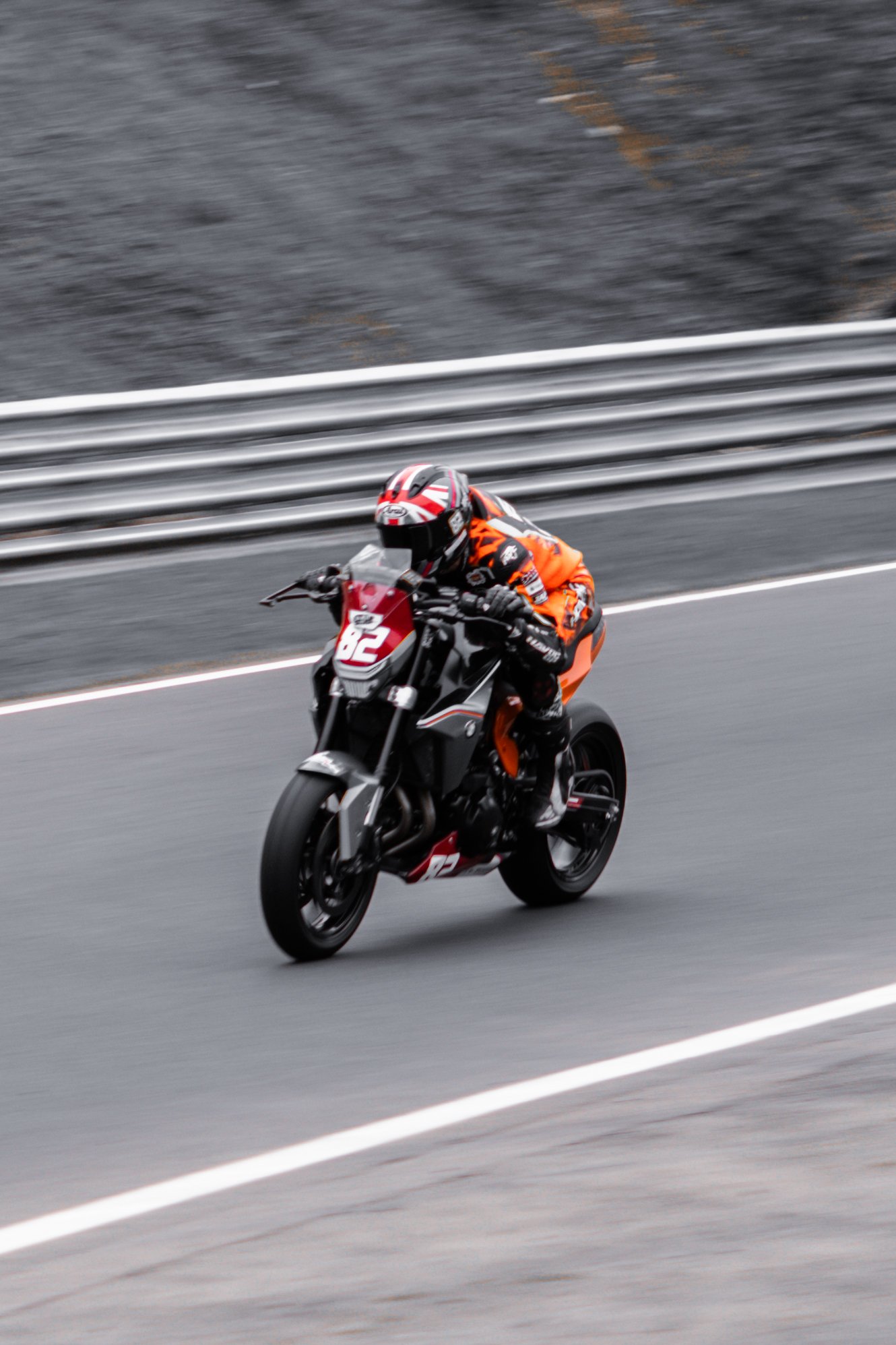 Motorcycle racer in orange and black gear riding a black and orange motorcycle on a racetrack, with gray barriers in the background.