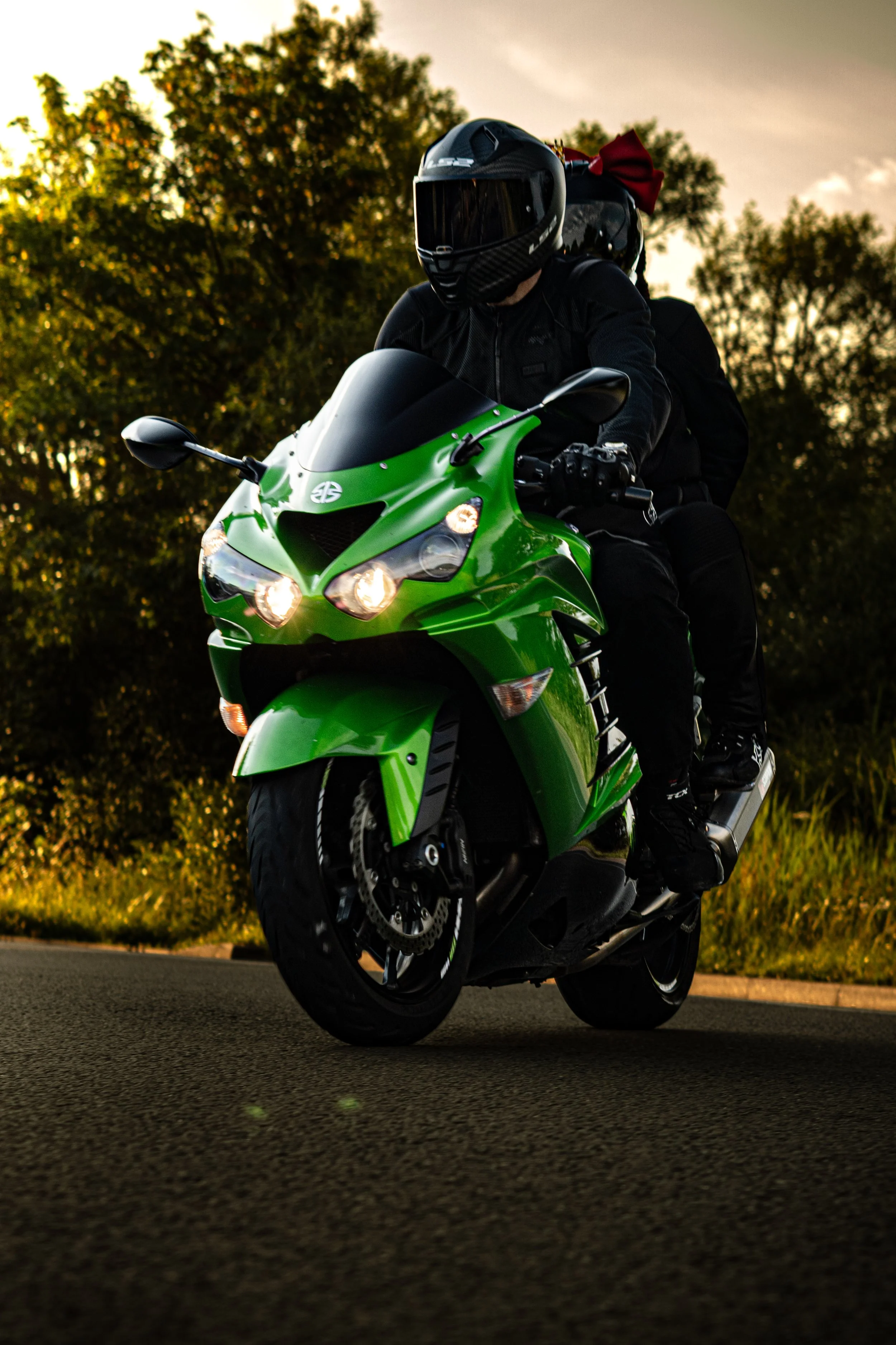 A person wearing a black helmet and riding a bright green motorcycle on a road with trees and a sunset sky in the background.