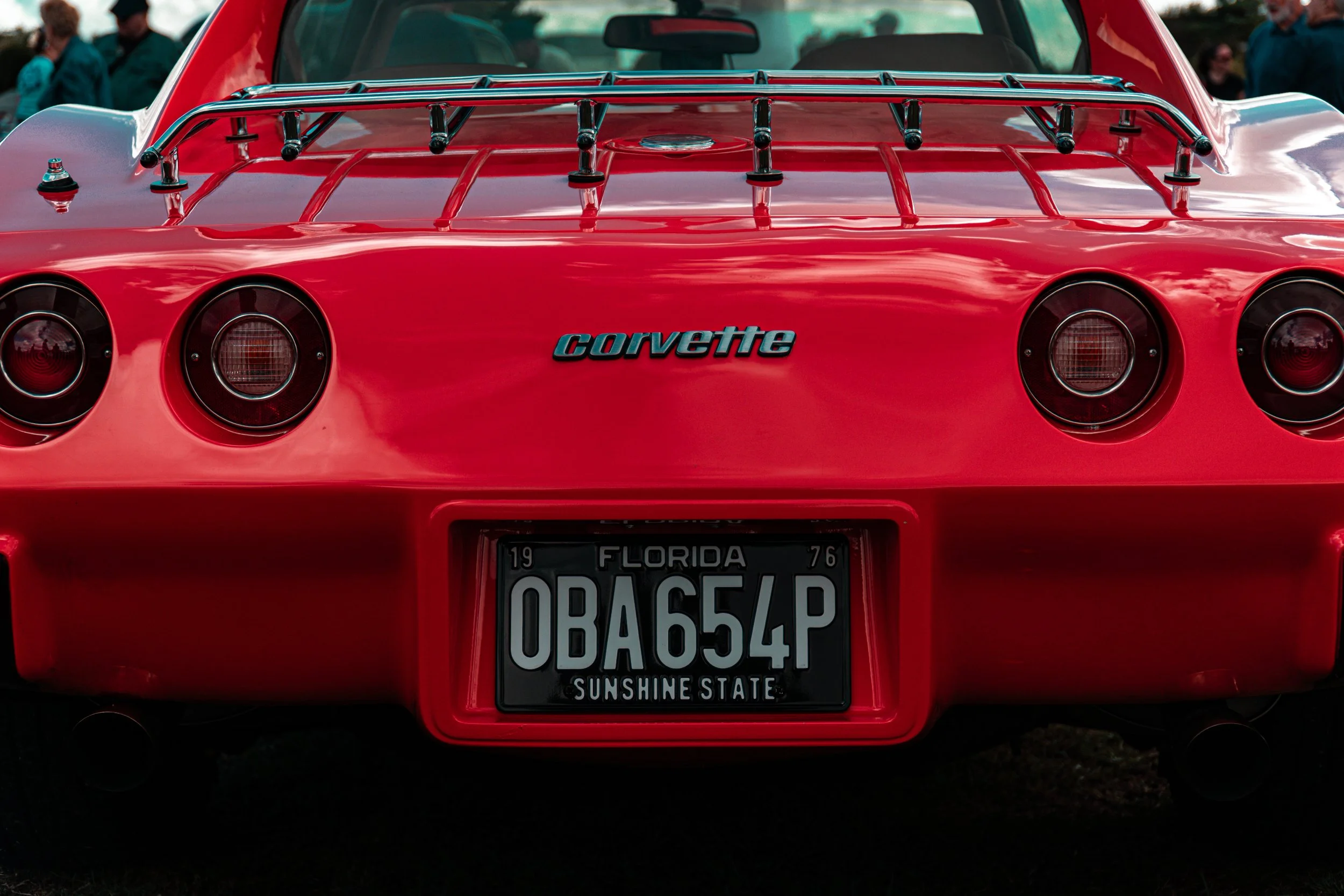 Rear view of a red Chevrolet Corvette sports car with a license plate from Florida that reads 'OBAGED4P' and a custom 'Corvette' badge on the back, with some people in the background.