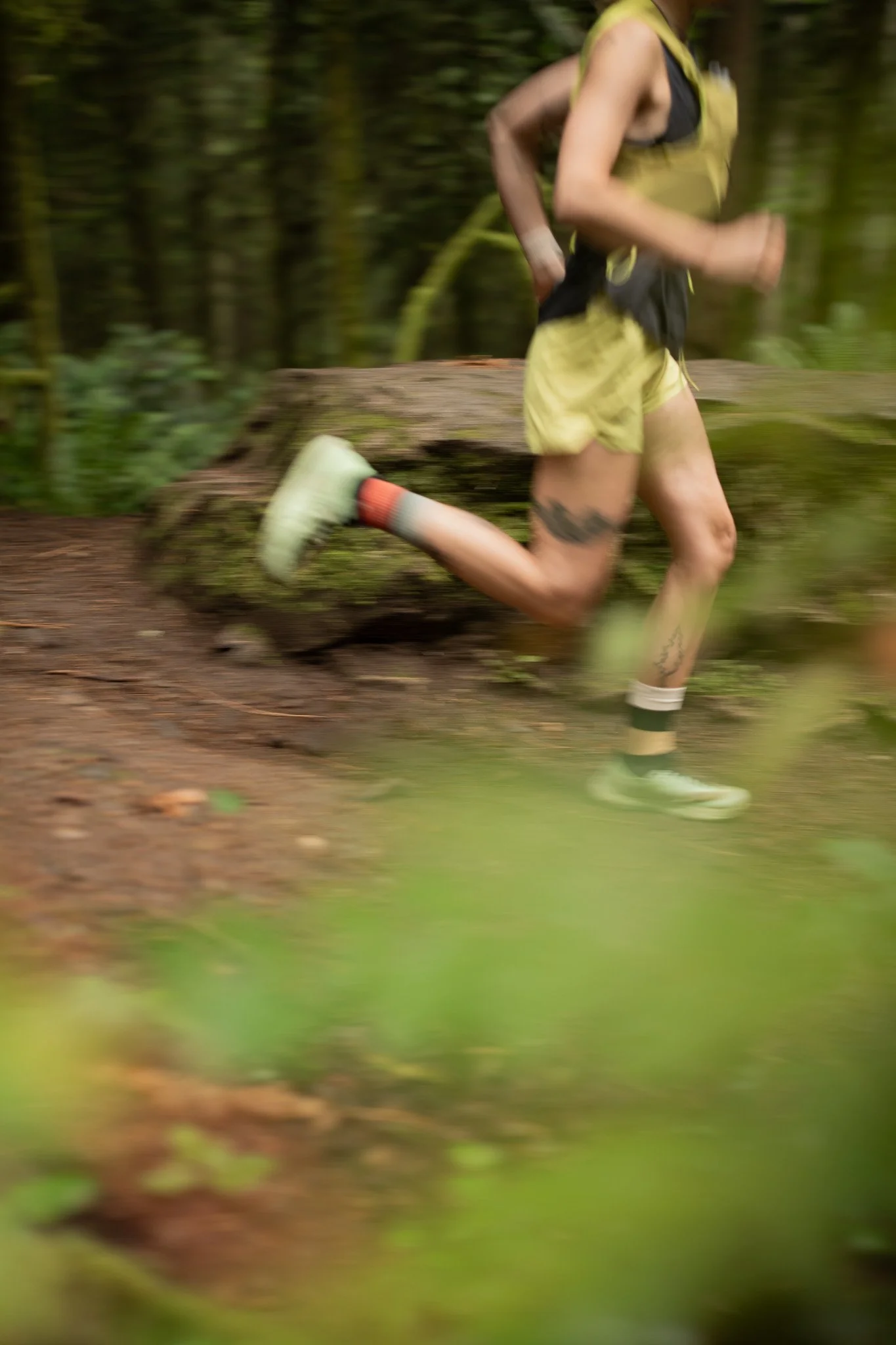 A person running on a forest trail, wearing yellow athletic gear, with tattoos visible on their legs. The background is a lush green forest.
