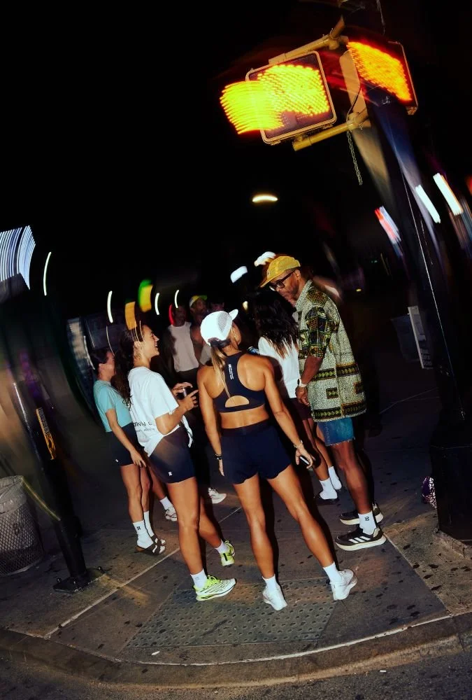 Group of young women and one man standing at a street corner at night, with a flashing yellow pedestrian crossing signal overhead.