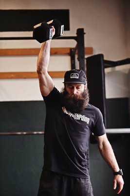 A man with a beard lifting a dumbbell overhead in a gym.