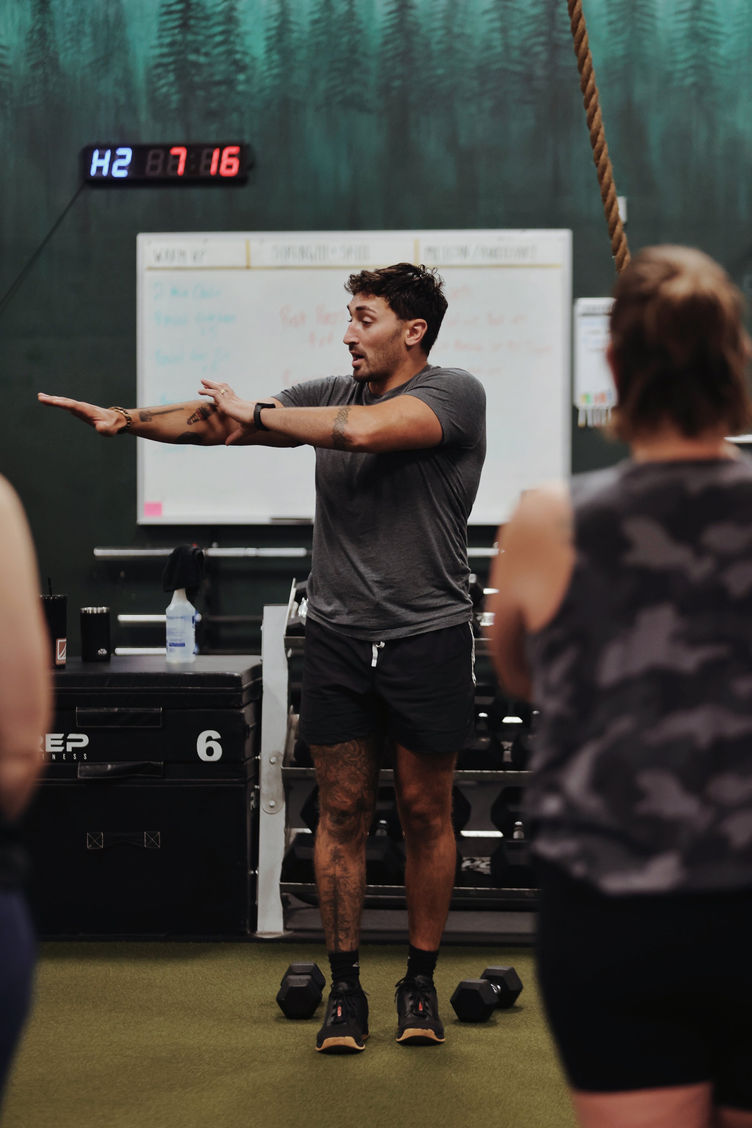 A fitness coach demonstrating an exercise in a gym classroom with a whiteboard, kettlebells, and a digital clock on the wall.
