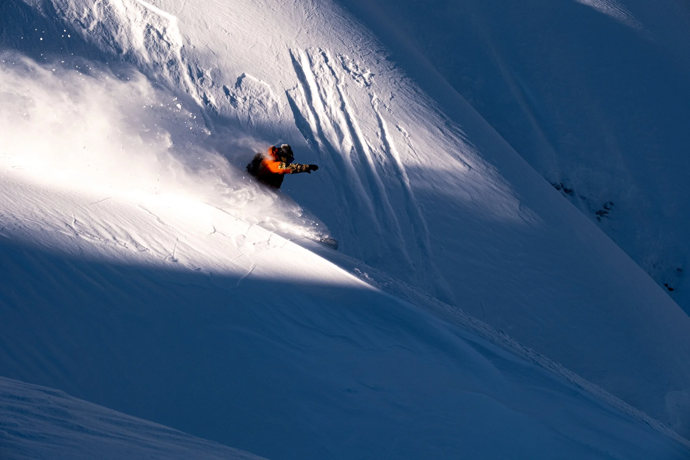 A skier in orange and black gear skiing down a snowy mountain slope, creating a spray of snow as they navigate the steep terrain under sunlight.