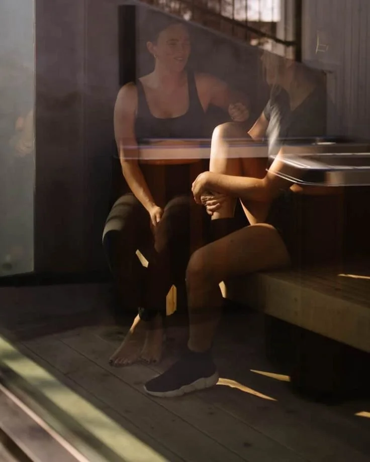 Two women sitting across from each other at a table, seen through a glass window with reflections. One woman is smiling, wearing a sleeveless top, while the other woman, with short hair, is dressed in athletic wear and is sitting with her legs crossed.