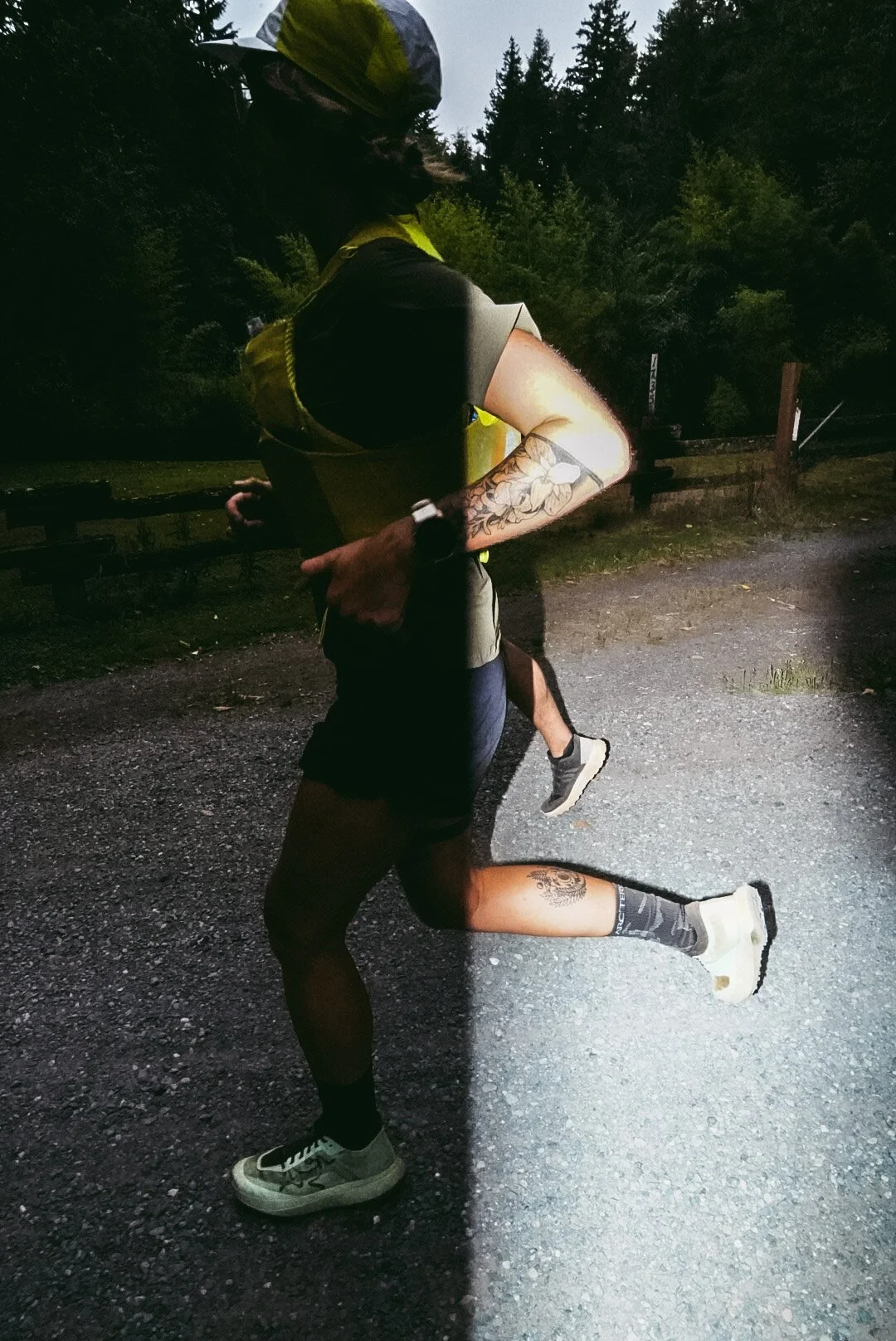 A person running outdoors at dusk, wearing athletic clothing and a cap, with visible tattoos on arms and legs, illuminated by a flashlight or headlamp.