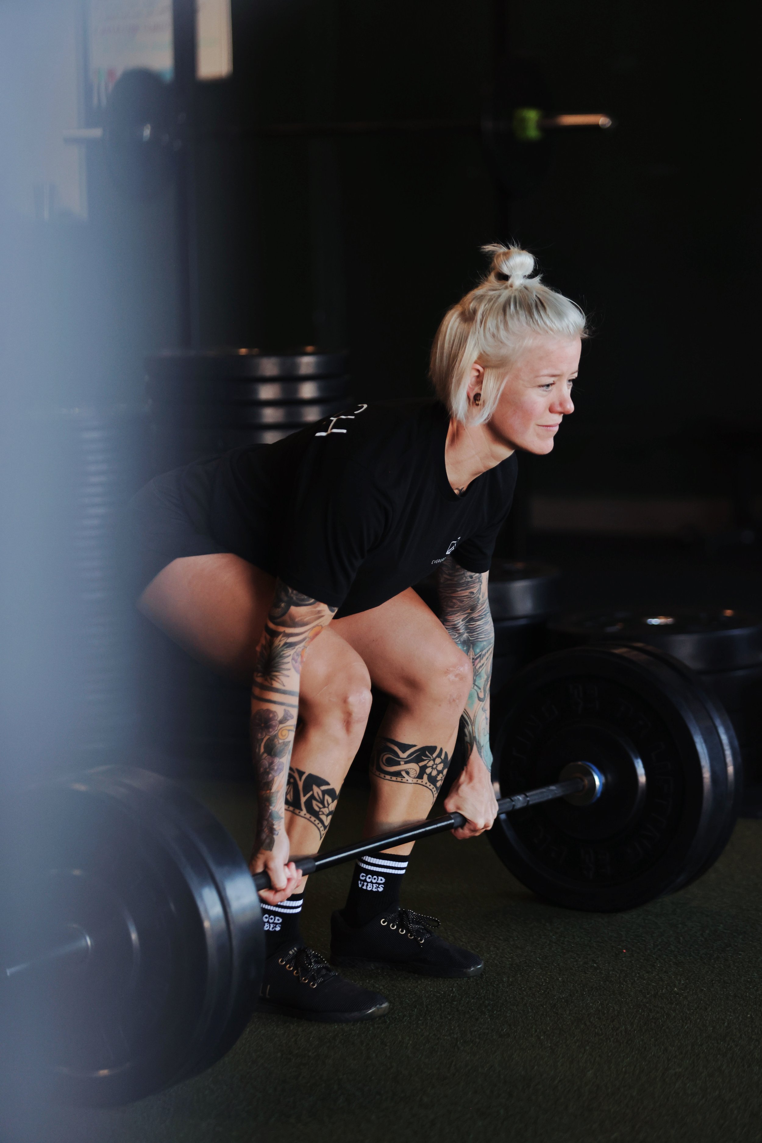 A woman with tattoos and blonde hair tied in a bun performing a deadlift with a barbell in a gym.