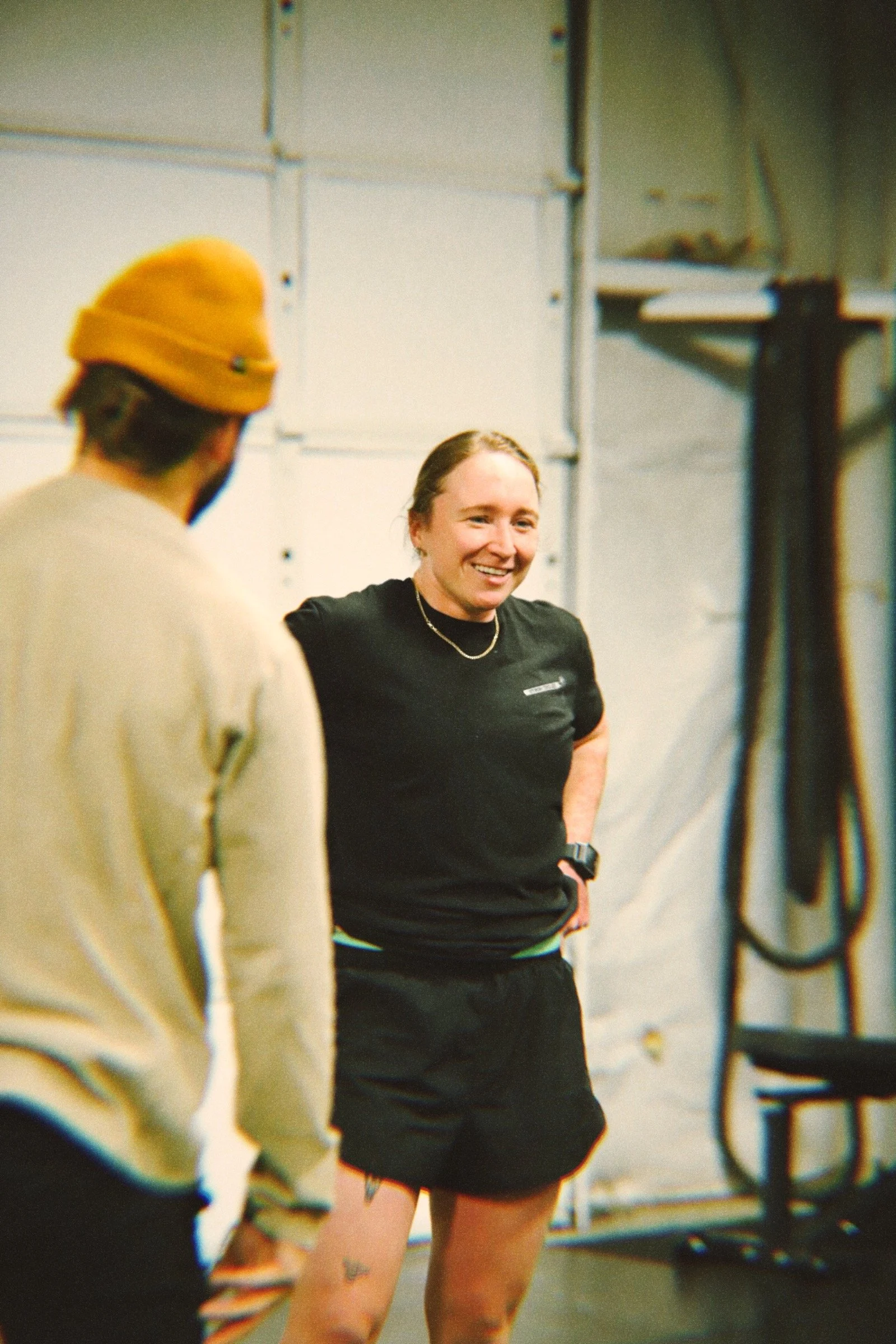 Two people talking in a gym. One woman, smiling, wearing a black t-shirt and black shorts. One man, with a yellow beanie and beige sweater, facing away from the camera.