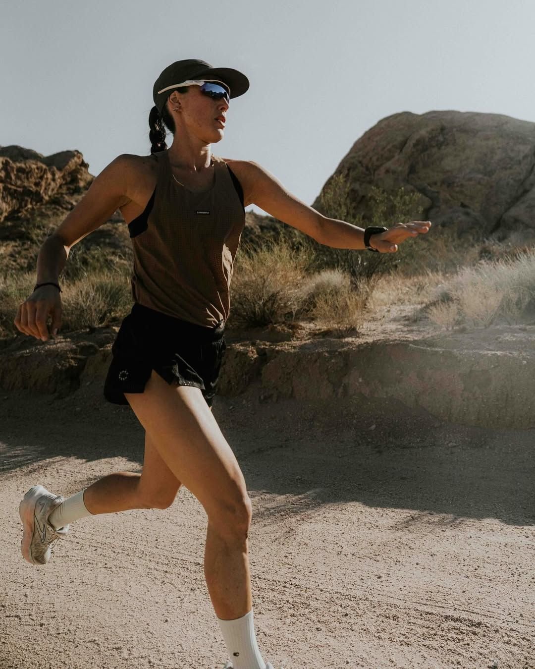 A woman running in a desert landscape with rocky formations in the background, wearing sunglasses, a hat, tank top, shorts, and running shoes.