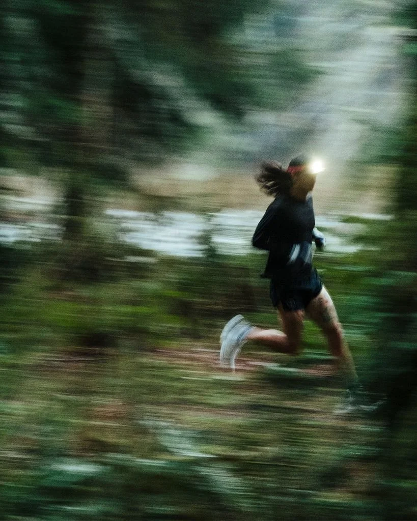 A woman running through a forested trail wearing a headlamp, black athletic clothing, and running shoes, captured with motion blur.
