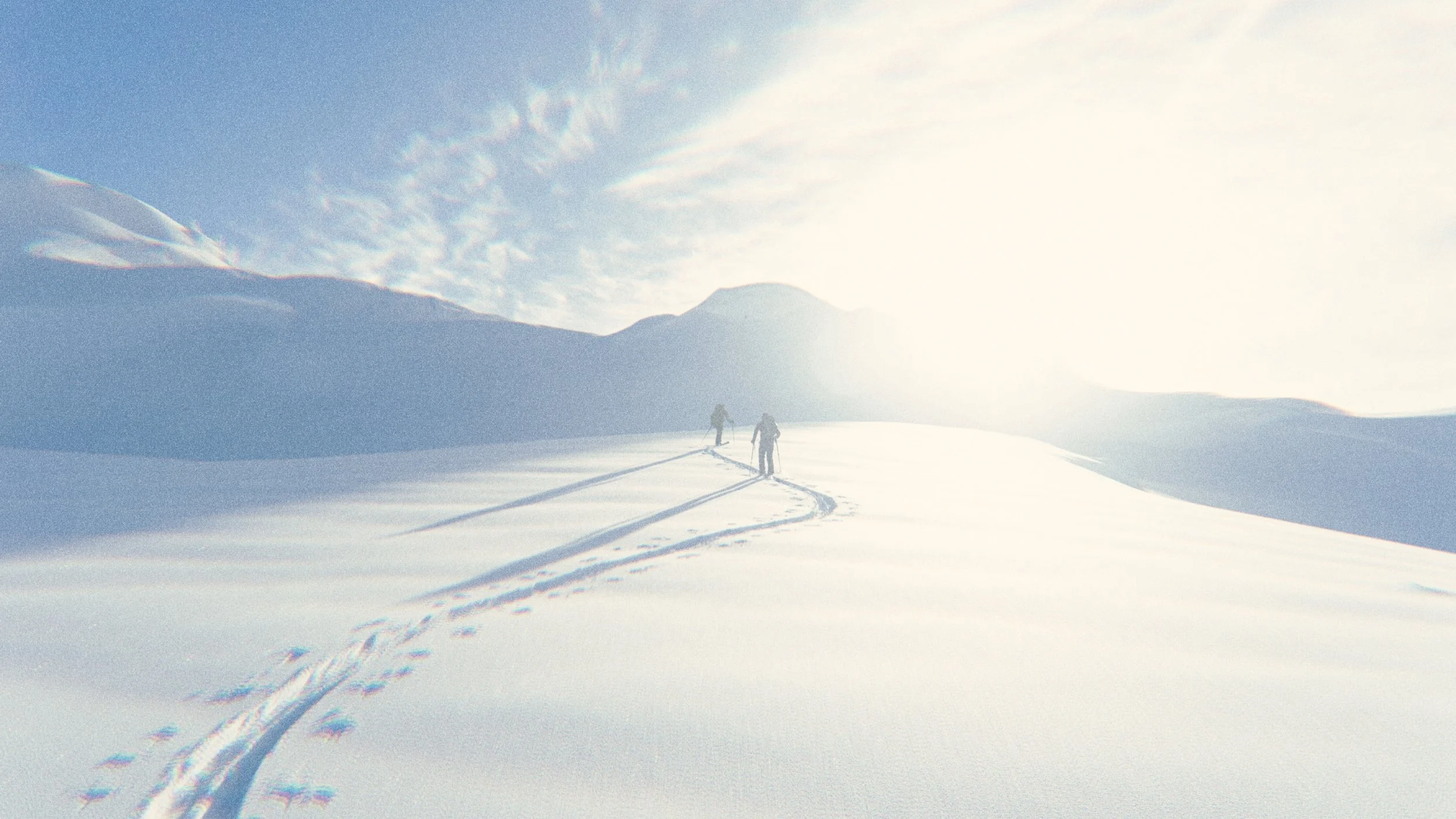 Two people hiking on snowy mountain trail with footprints, under bright sunlight and partly cloudy sky.