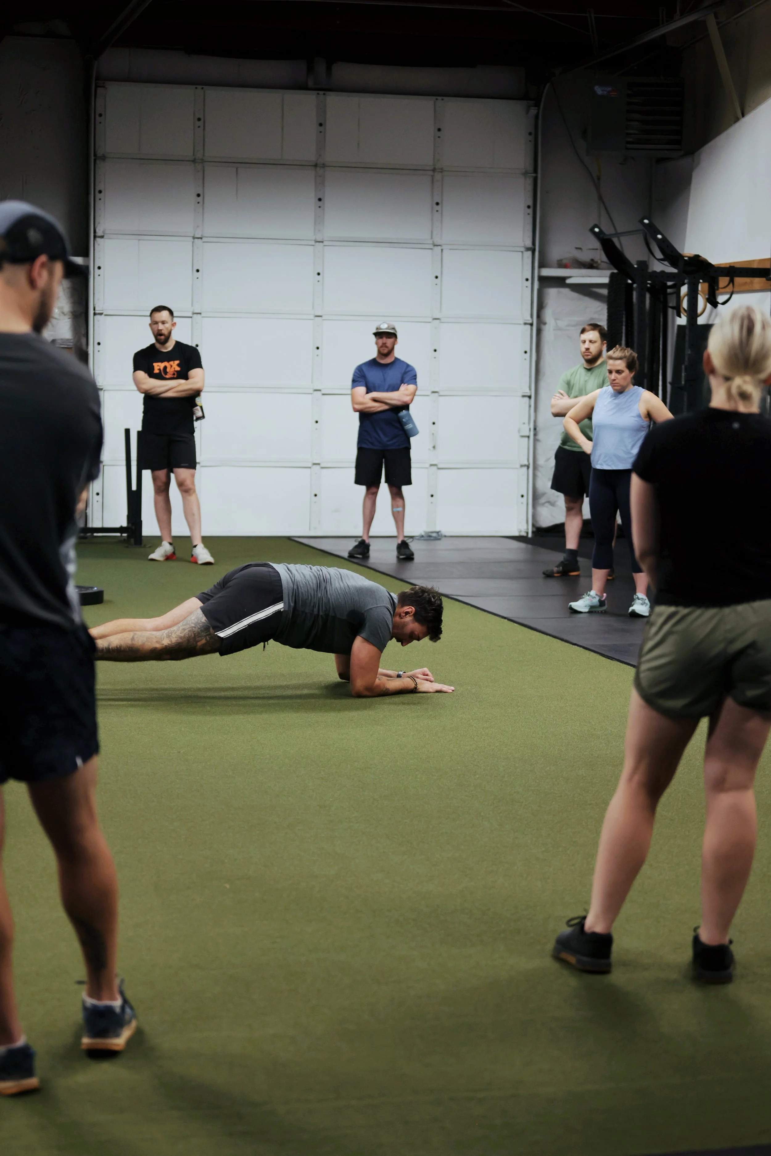 A group of people at a gym, with one man performing a plank exercise on the green turf while others watch, some with crossed arms.