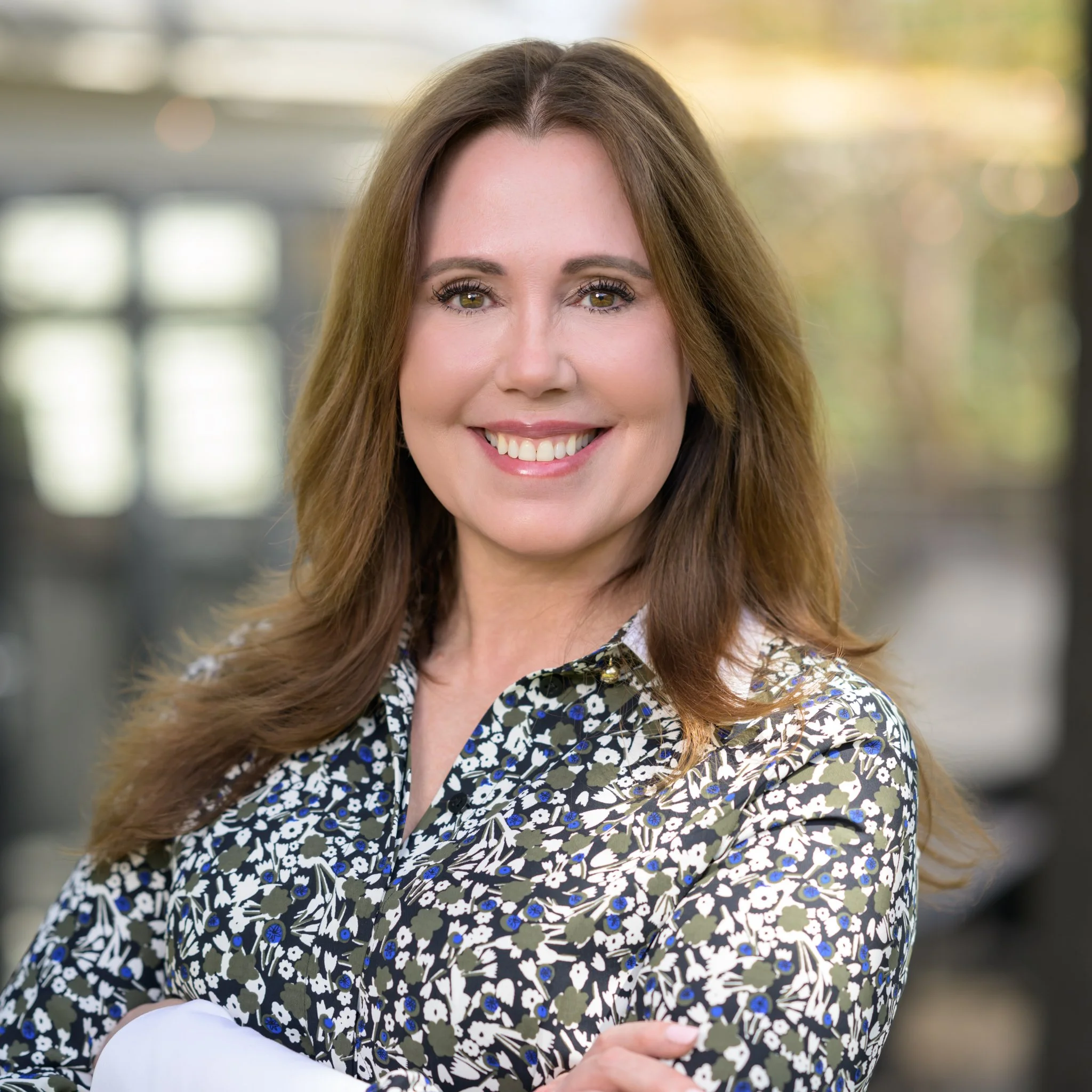 Smiling woman with long, wavy brown hair wearing a floral blouse standing outdoors with blurred trees and buildings in the background.