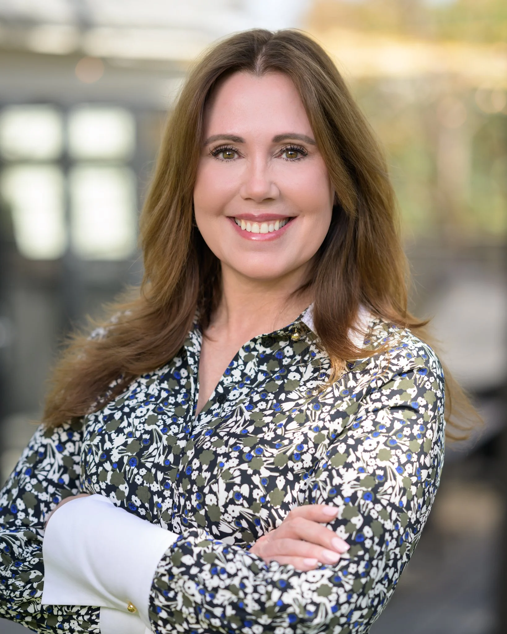 Smiling woman with long light brown hair, wearing a floral patterned blouse, crossing her arms, in an indoor setting with blurred background.