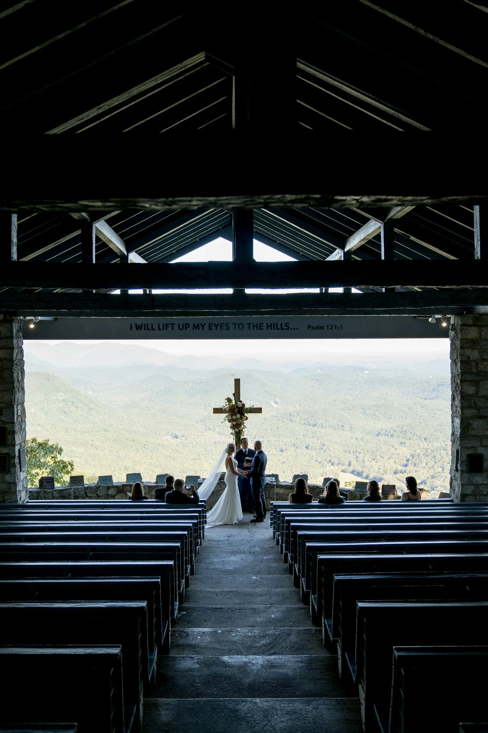A wedding ceremony inside a chapel overlooking mountains, with a bride and groom holding hands at the altar, guests seated in pews, a cross with floral arrangement behind them, and a sign with a Bible verse.