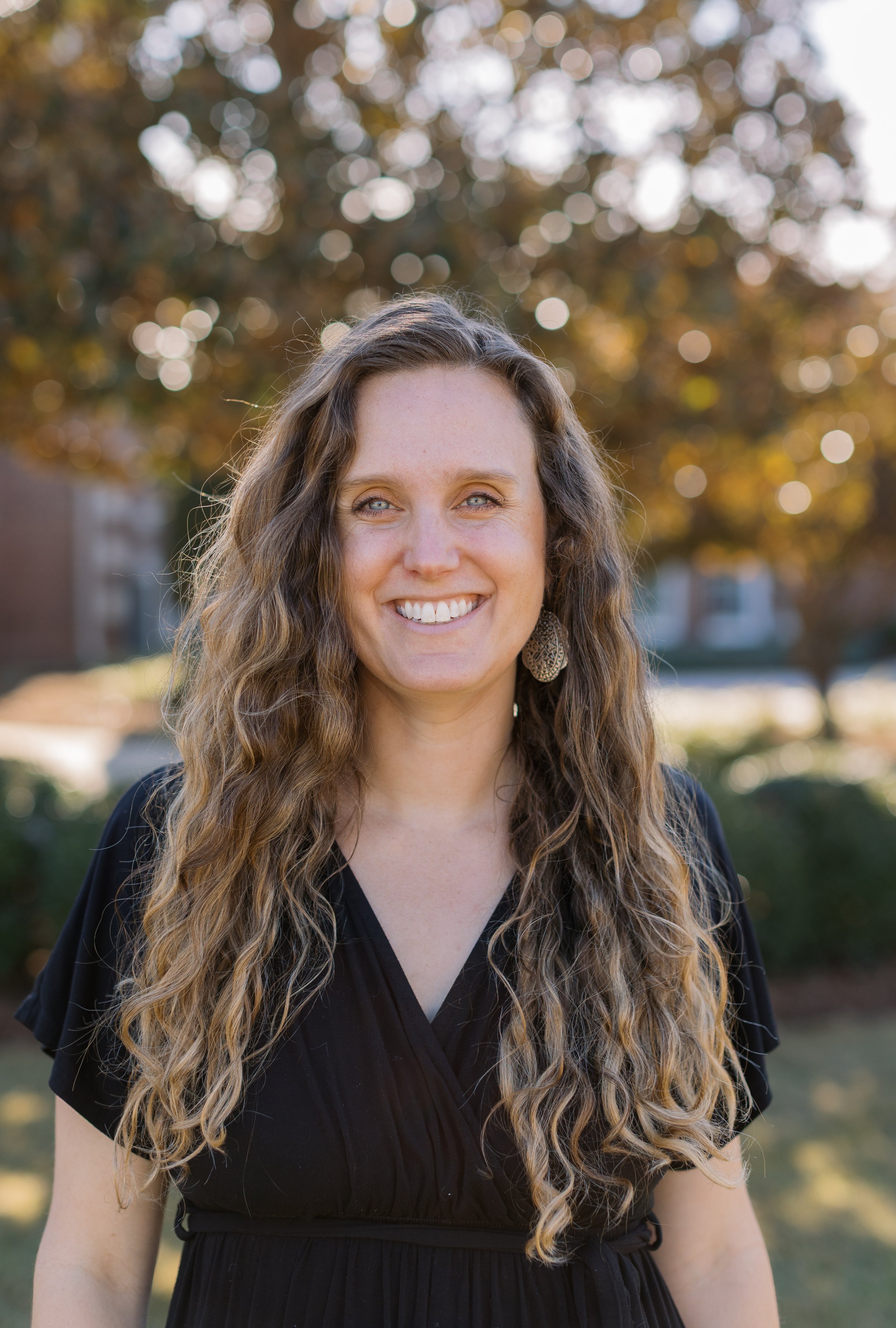 A woman with long, curly brown hair and blue eyes smiling outdoors on a sunny day with trees and houses in the background.