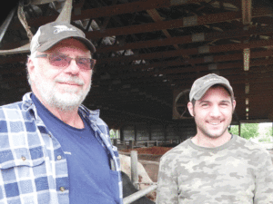 Two men standing outdoors near a wooden structure. One is older with a beard, wearing glasses, a cap, a blue plaid shirt, and a navy t-shirt. The other is younger with a beard, wearing a cap and camouflage shirt.