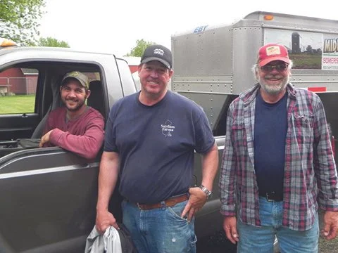 Three men standing outdoors beside a pickup truck and a trailer, smiling at the camera.