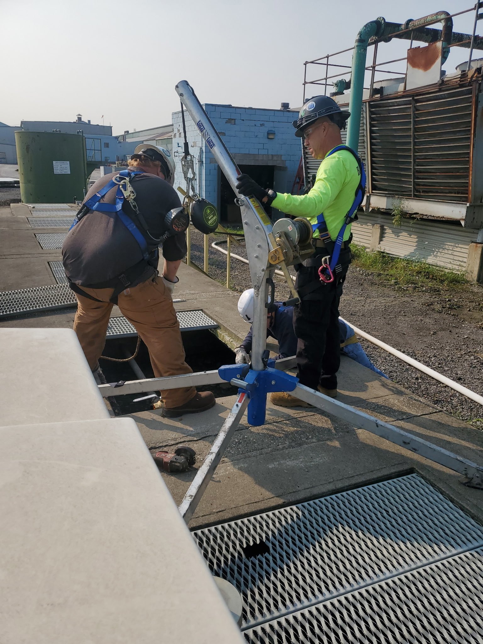 Two workers in safety gear, including helmets and harnesses, are working at a site with industrial equipment and pipes outdoors. One worker is bending over, and the other is standing, holding a tool near a tripod-like device.