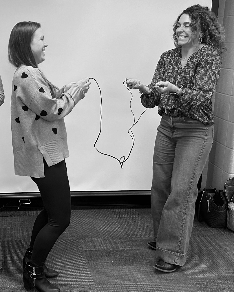 Two women are smiling and laughing while passing a jump rope to each other indoors.