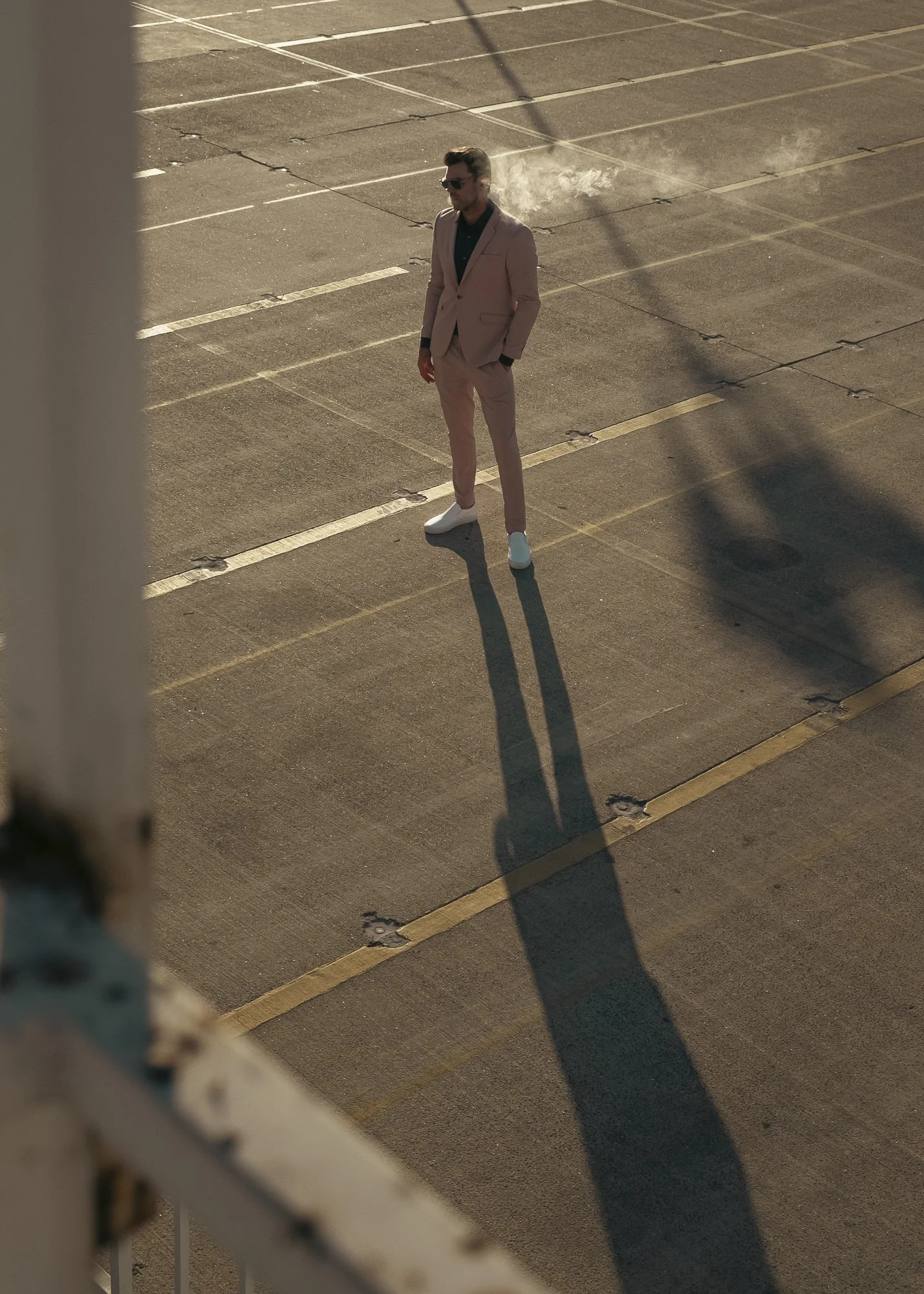 A man in a light pink suit, black shirt, and white shoes standing in an empty parking lot, casting a long shadow and smoking, with sunlight creating shadows.