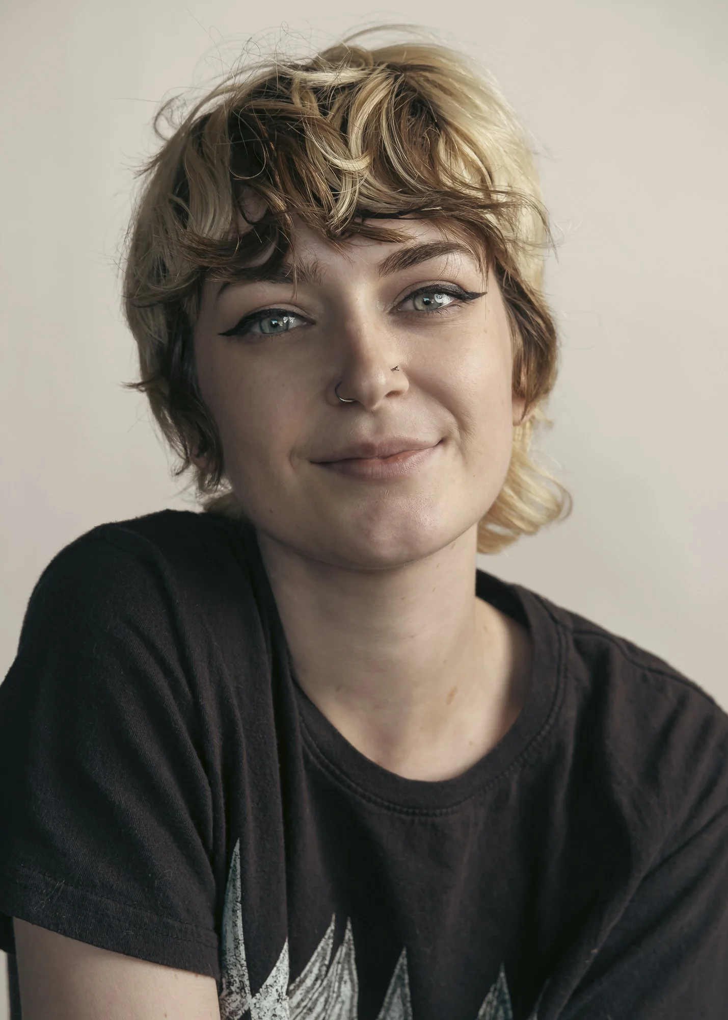 A woman with short, curly, blonde hair, wearing black eyeliner and a black T-shirt, smiling softly at the camera against a plain light-colored background.
