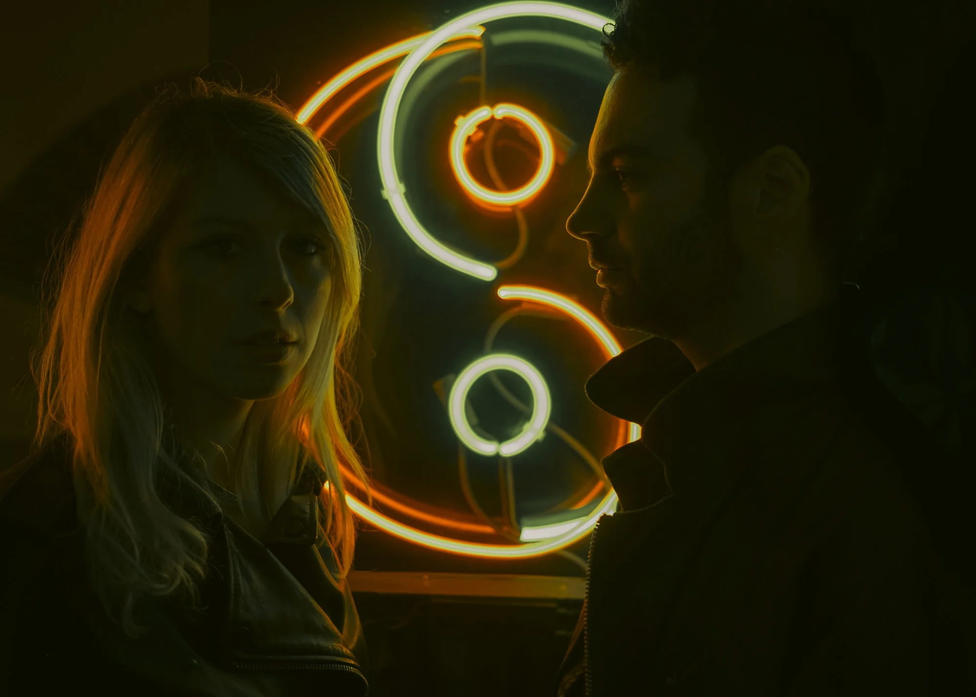 A woman and a man are facing each other in front of a neon light art installation with green and orange glowing circles and swirls, creating a vibrant, abstract background.