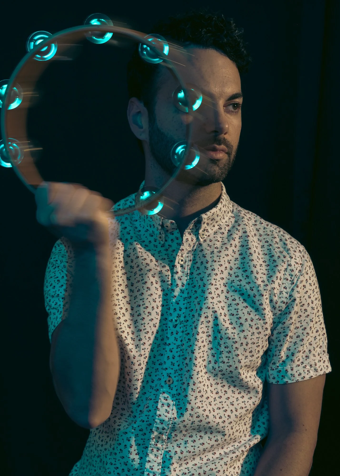 A man with dark, curly hair and a beard, holding a circular device emitting blue lights, against a dark background. He is wearing a patterned button-up shirt.