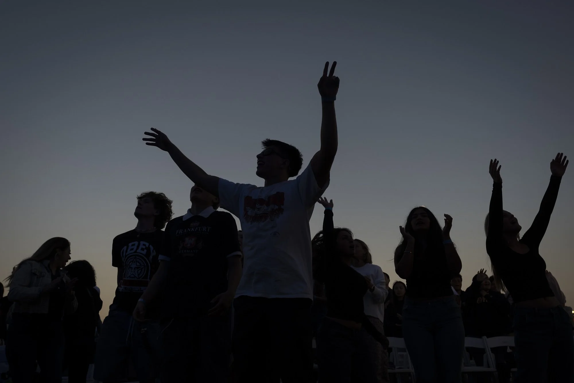 Silhouettes of people at sunset, some with arms raised, in an outdoor gathering.