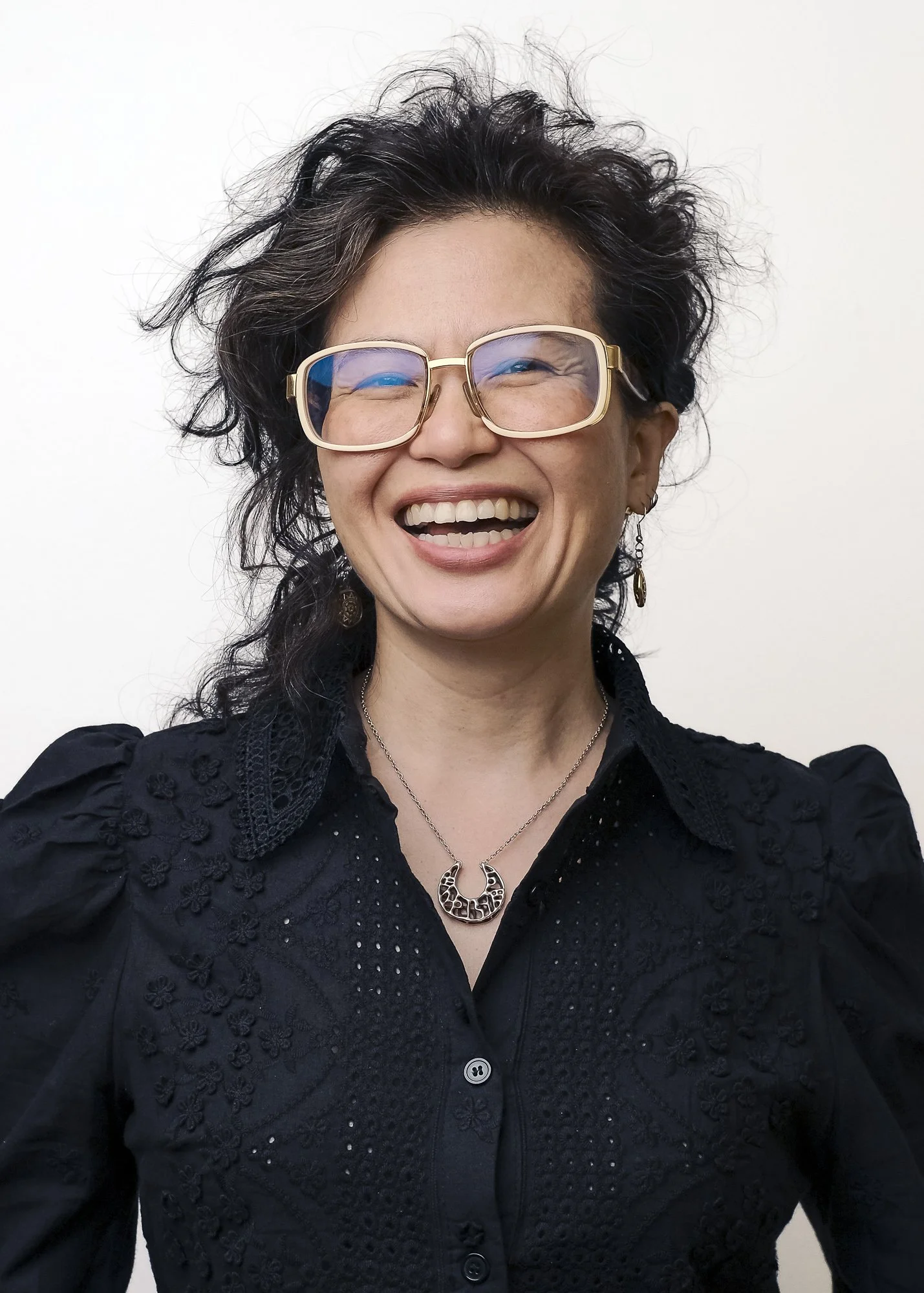 A woman with curly dark hair and glasses smiling, wearing a black embroidered blouse and jewelry.