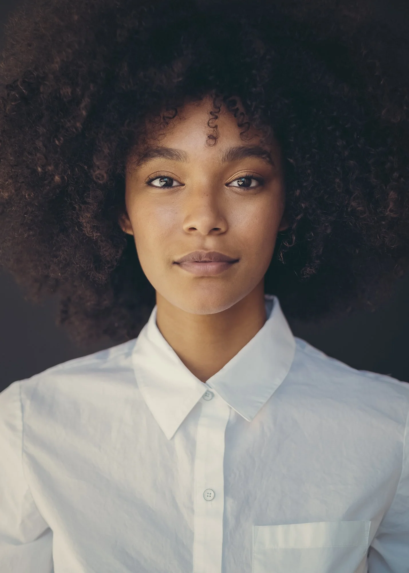 Close-up portrait of a young Black woman with a large, curly afro, wearing a white button-up shirt, looking directly at the camera with a calm expression.
