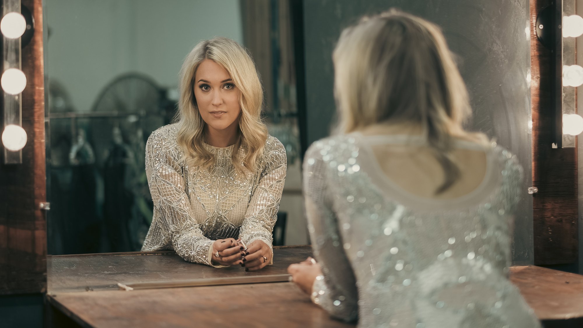 A woman with blonde hair looks at herself in a mirror with lights around it, sitting at a wooden dressing table.