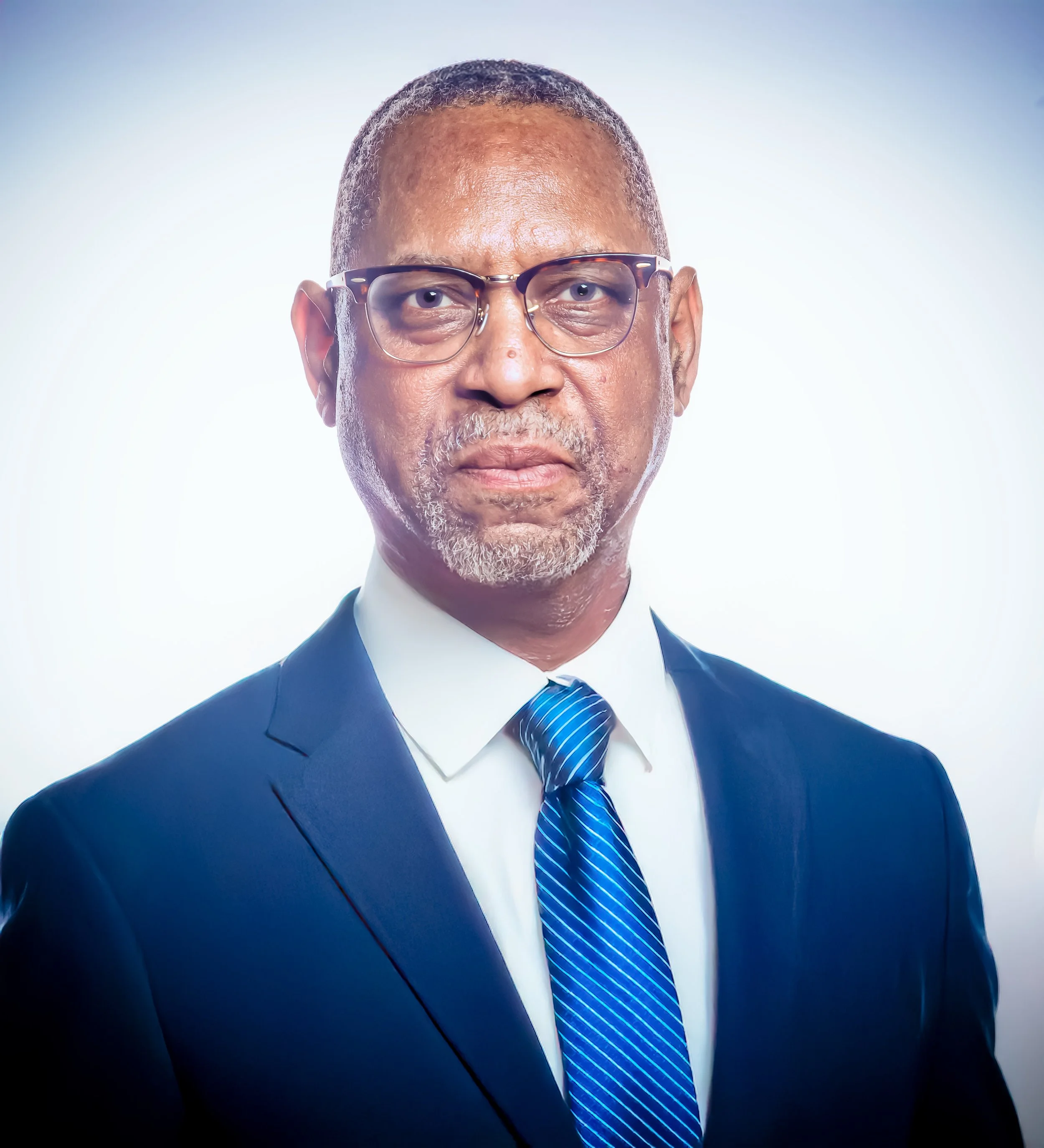 Close-up of a middle-aged man with black hair and glasses, wearing a dark blue suit, white shirt, and a blue striped tie, against a plain light background.