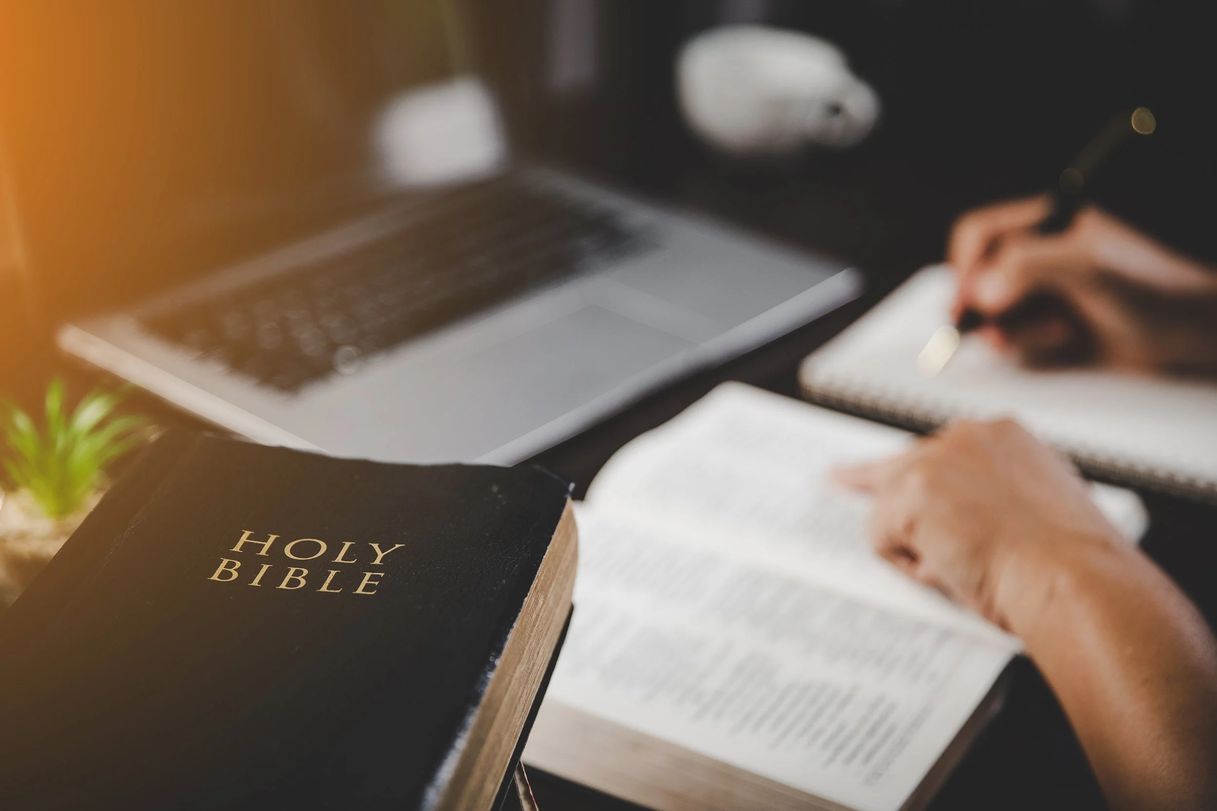 A person writing in a notebook with a stack of books, including a Holy Bible, and a laptop on a table.