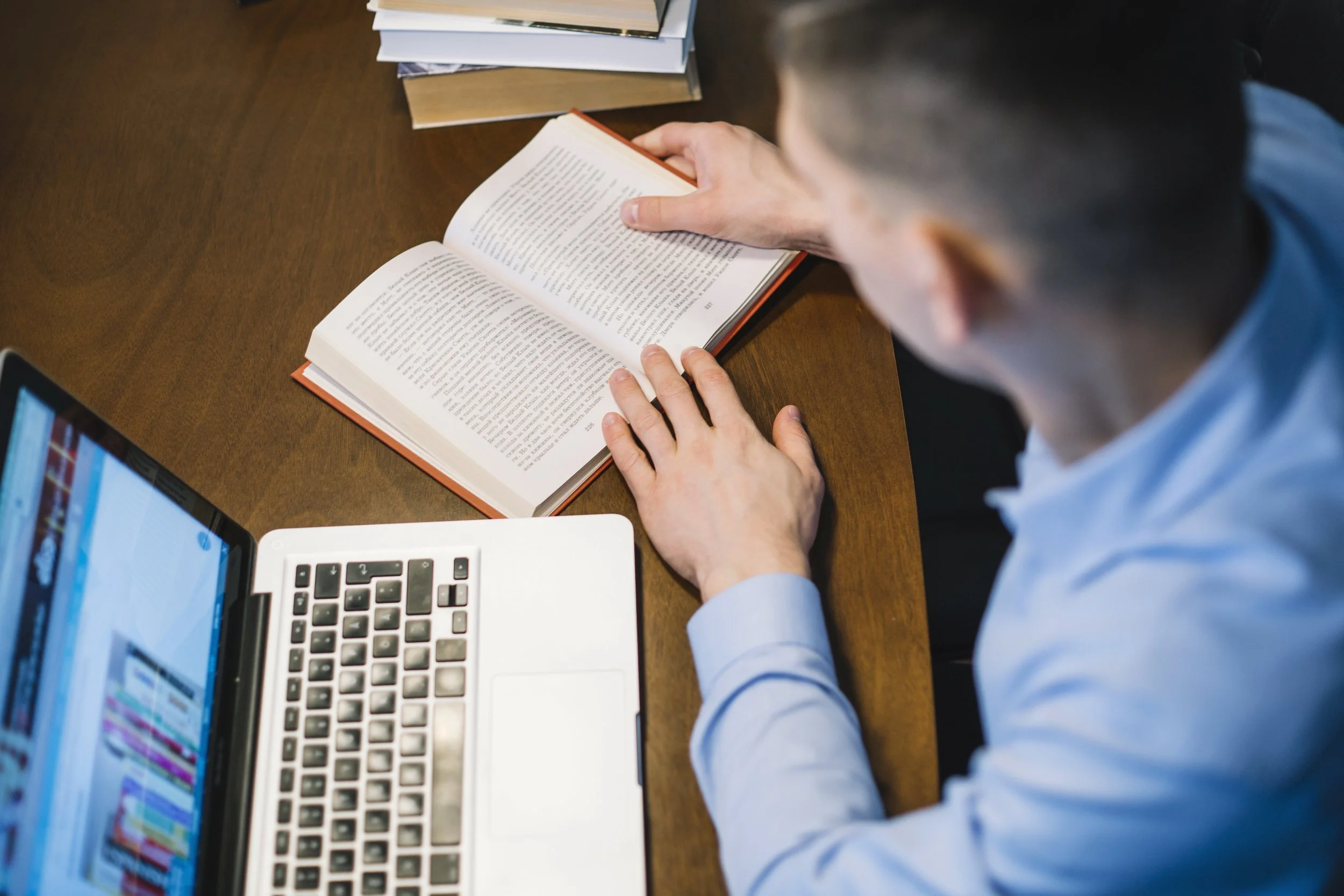 Person reading an open book at a desk with a laptop and a stack of books.