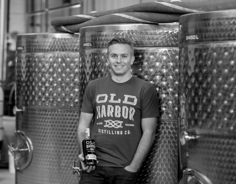 A smiling young man standing in front of large stainless steel tanks in a distillery, holding a bottle of Old Harbor Distilling Co. liquor. The tanks are used for brewing or distilling spirits.