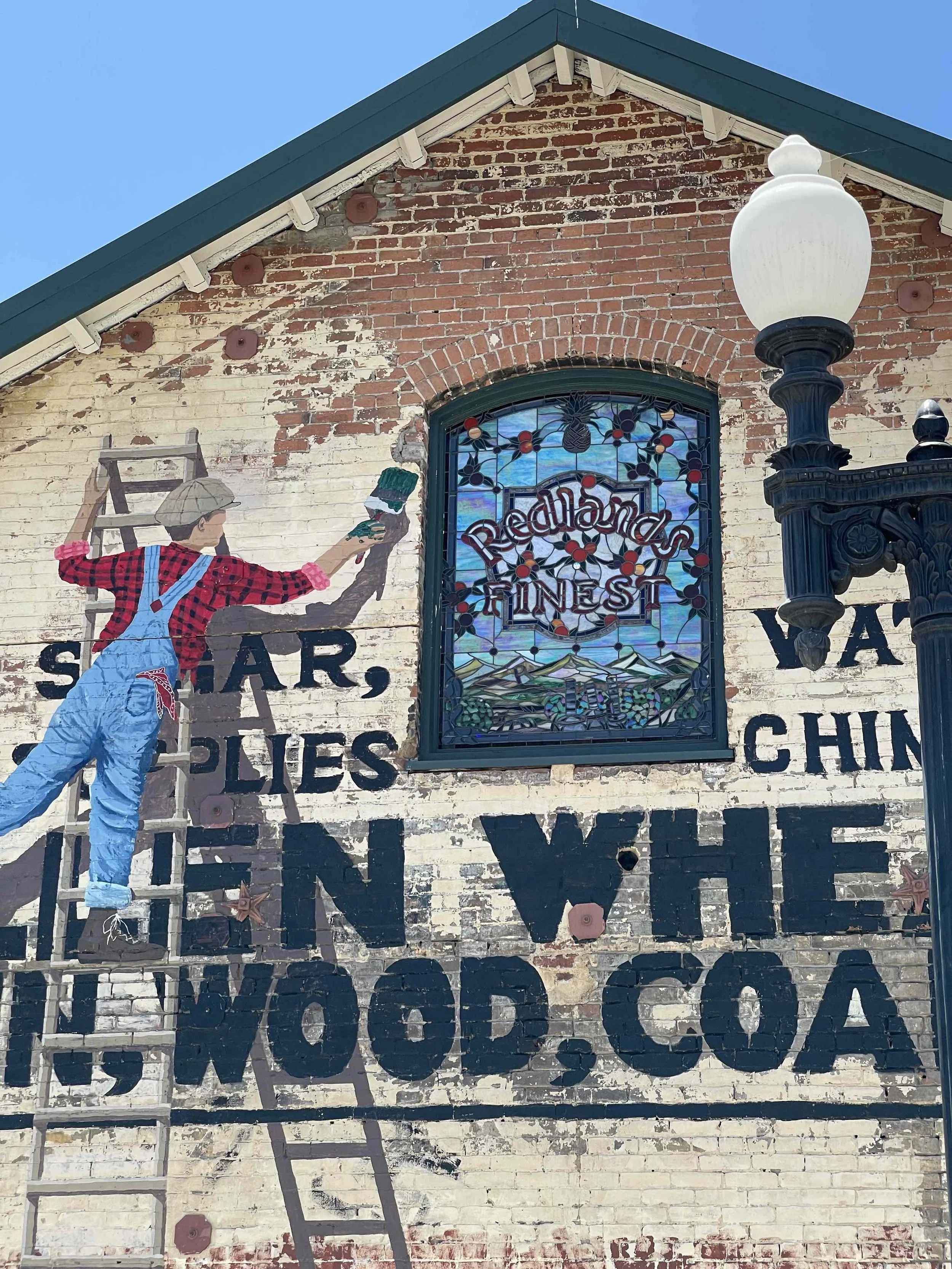 A mural on a brick building wall depicts a person in overalls and a hat painting a sign. The mural includes large black text and a stained-glass window that says 'Redland's Finest' with a scenic landscape. A black ornate street lamp is in front of th