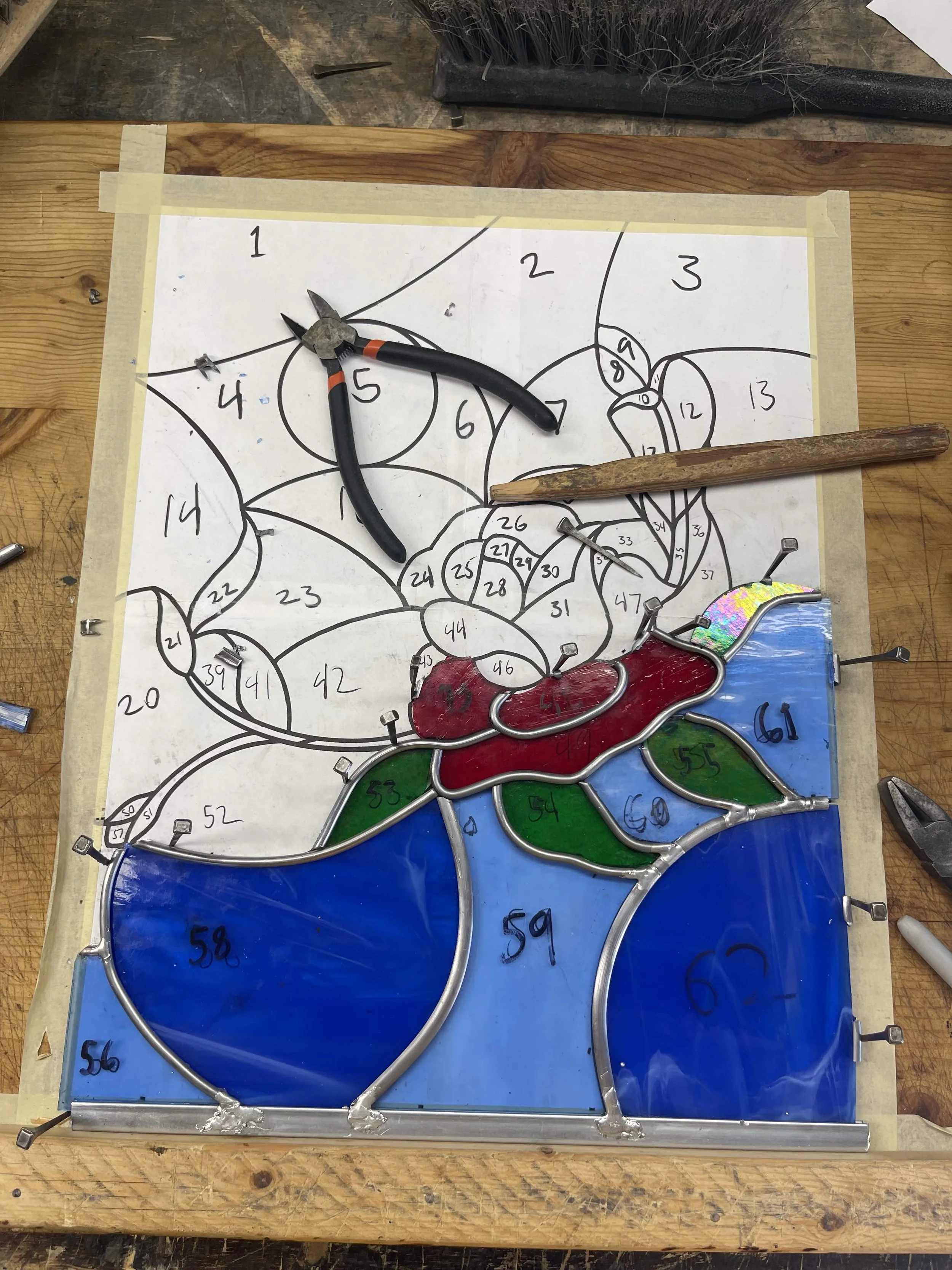 Colorful stained glass design in progress on a wooden workbench, with framing, tools, and a numbered pattern outline.