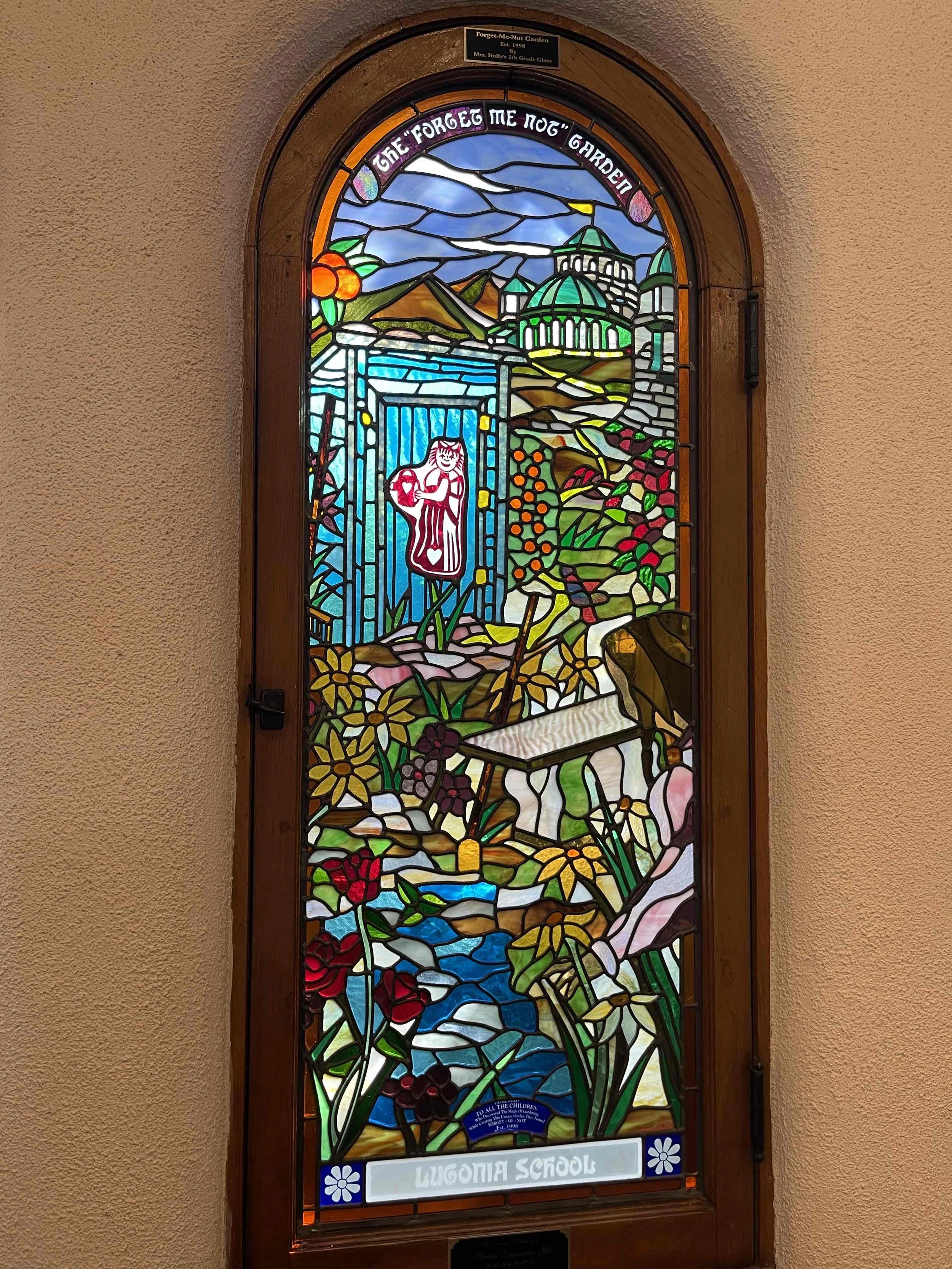 Colorful stained glass window depicting a garden scene with flowers, a stream, and a statue of a girl. The window has an arched top and is labeled with the words 'Lugonia School' at the bottom.