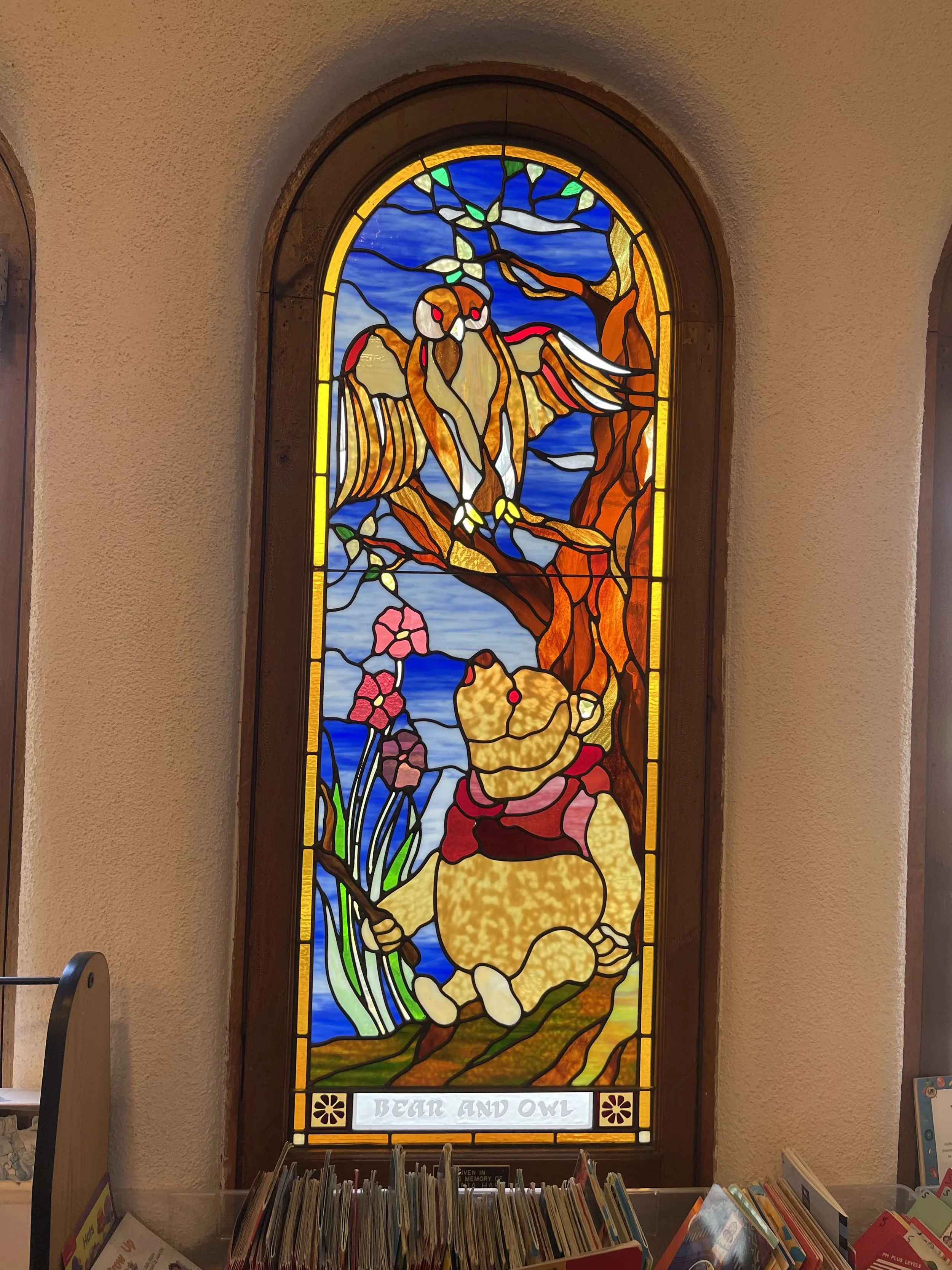 Colorful stained glass window depicting a bear sitting in a forest with an owl perched on a tree branch above. The window is labeled "Bear and Owl" at the bottom.