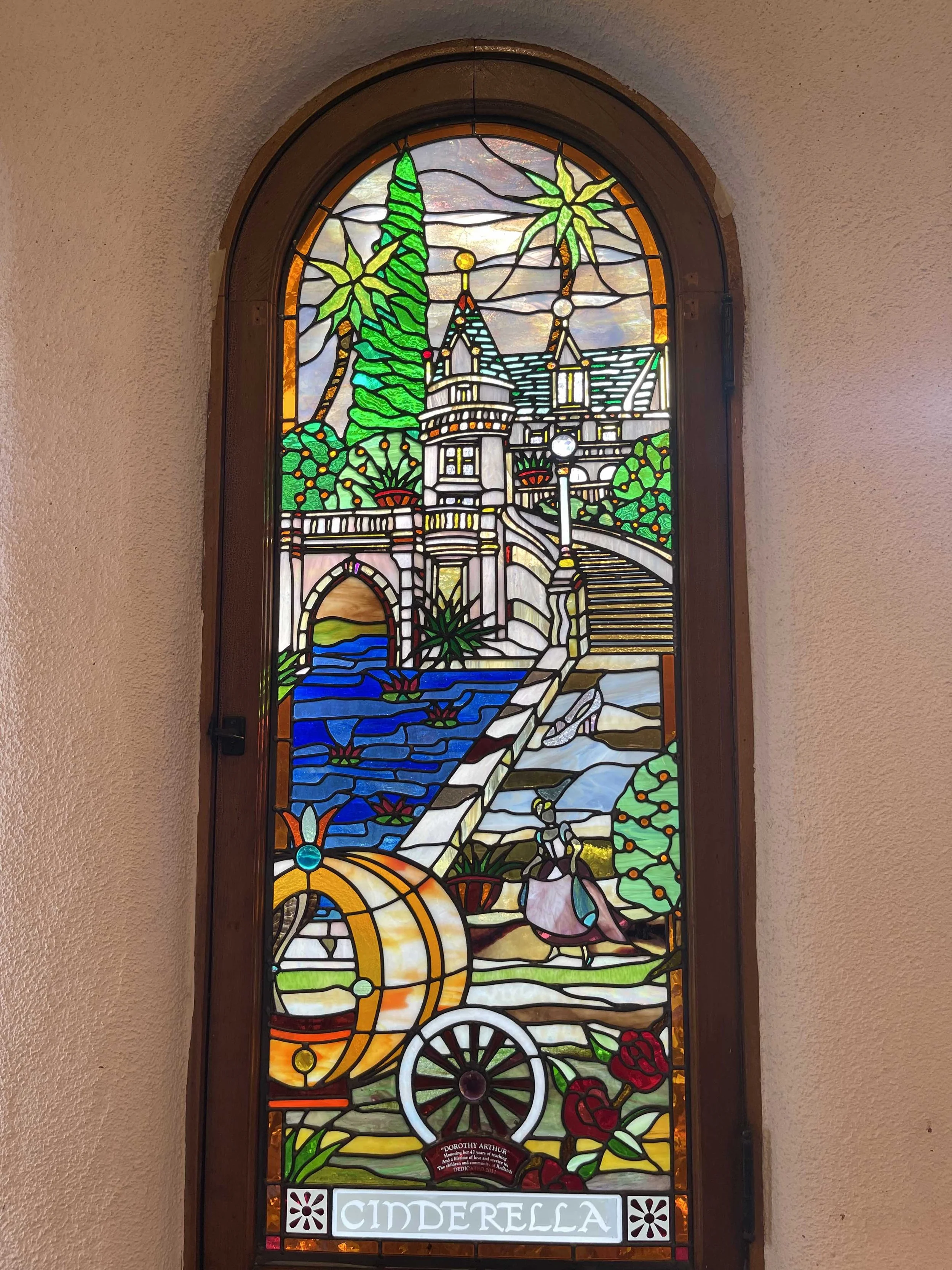 Stained glass window depicting Cinderella's pumpkin carriage, a castle, pathway, trees, and a fairy tale scene with the word 'CINDERELLA' at the bottom.