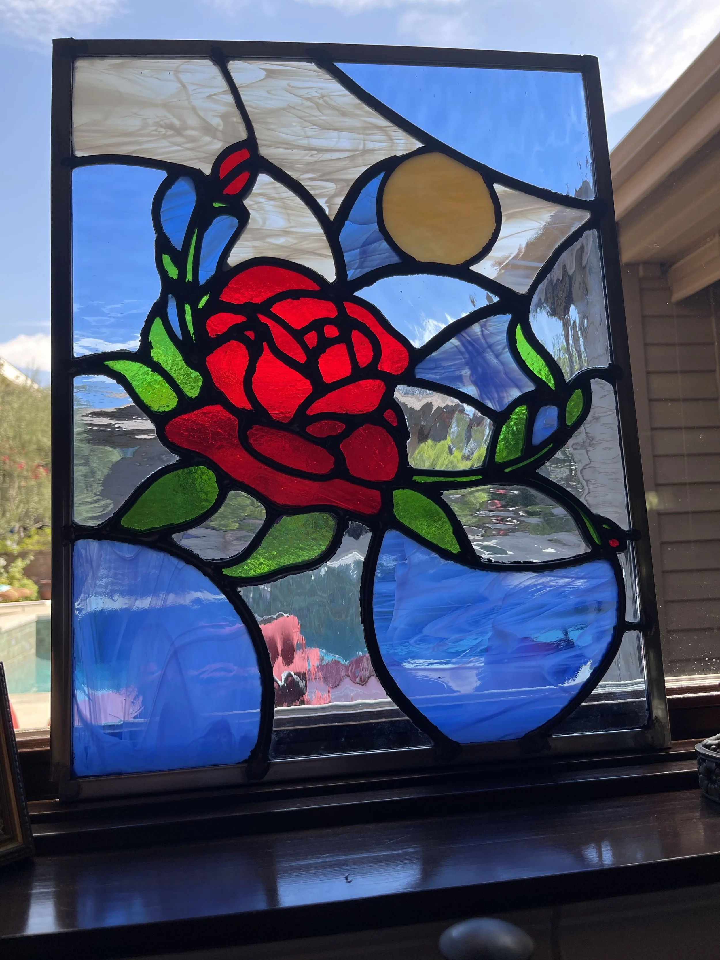 Stained glass window featuring a red rose, green leaves, a yellow sun, and blue sky elements.