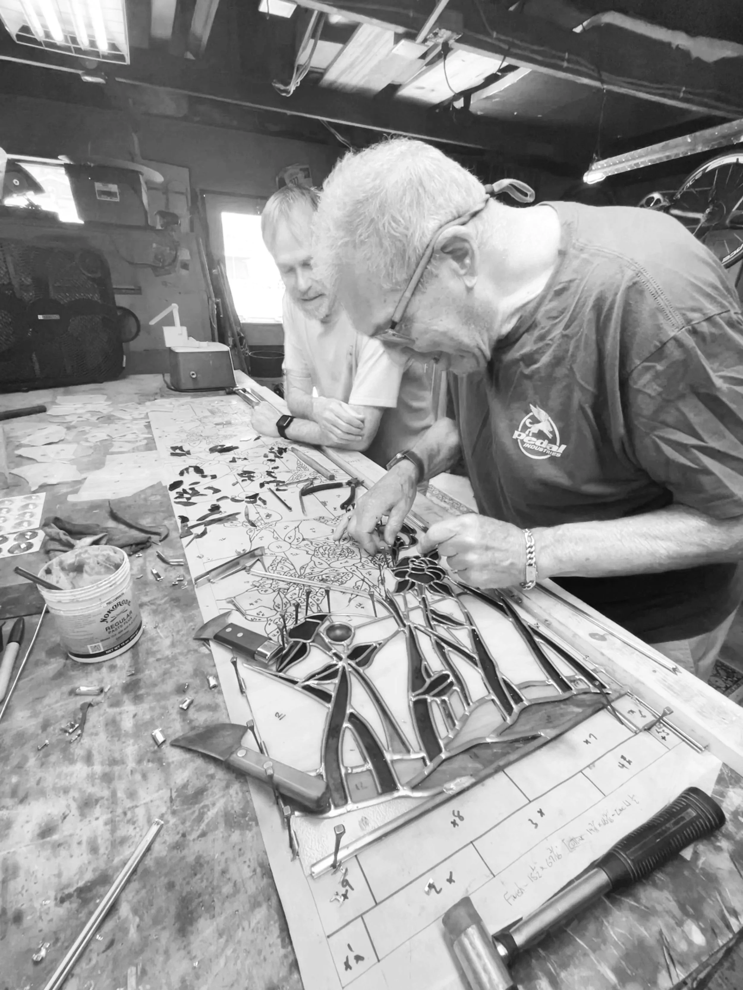 Two men working on stained glass art in a workshop, one assembling colored glass pieces while the other observes, with various tools and a work plan on the table.
