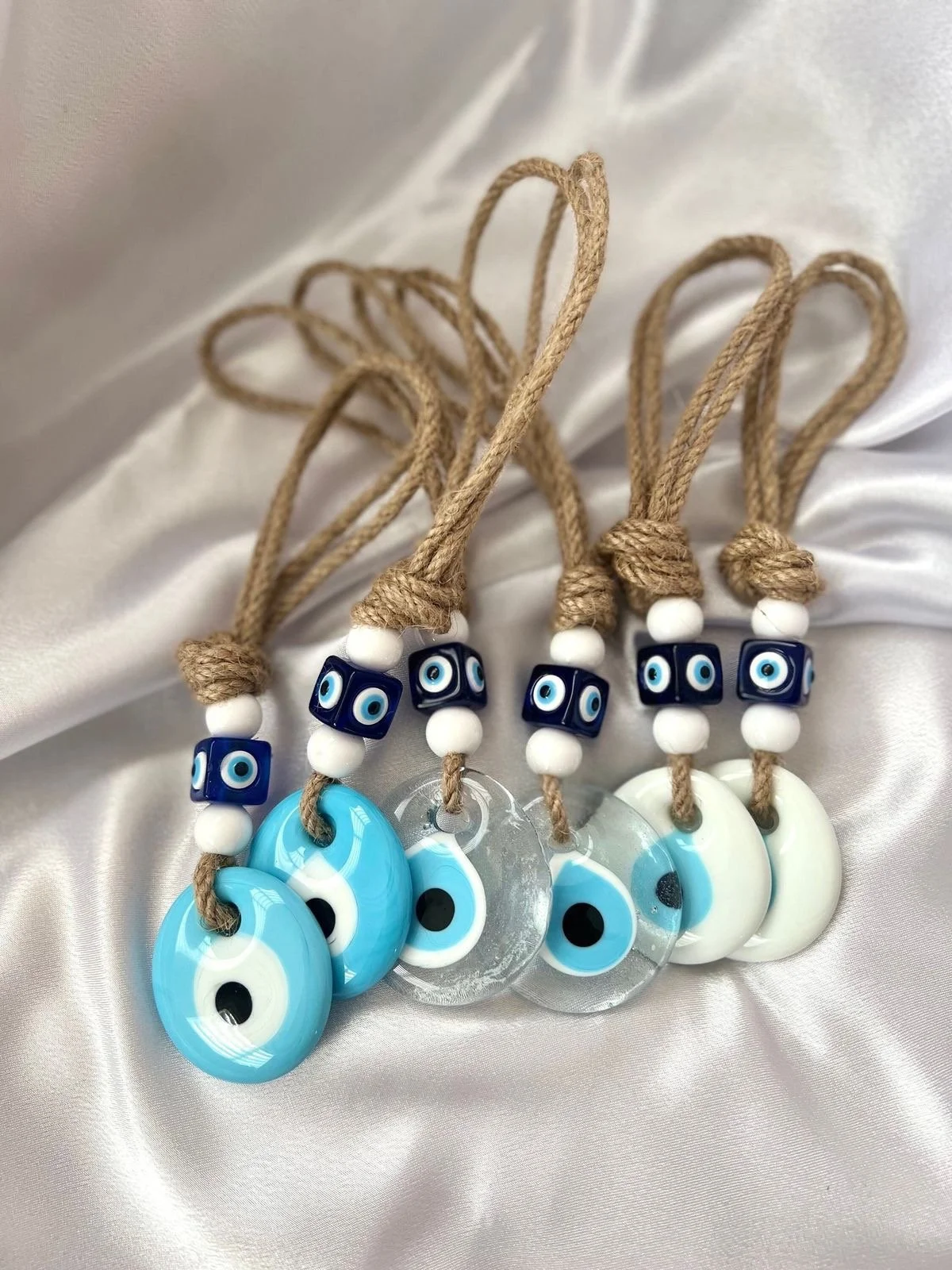 Set of five necklaces with blue eye charms, blue glass eye beads, white beads, and tan rope cords, displayed on a satin fabric background.