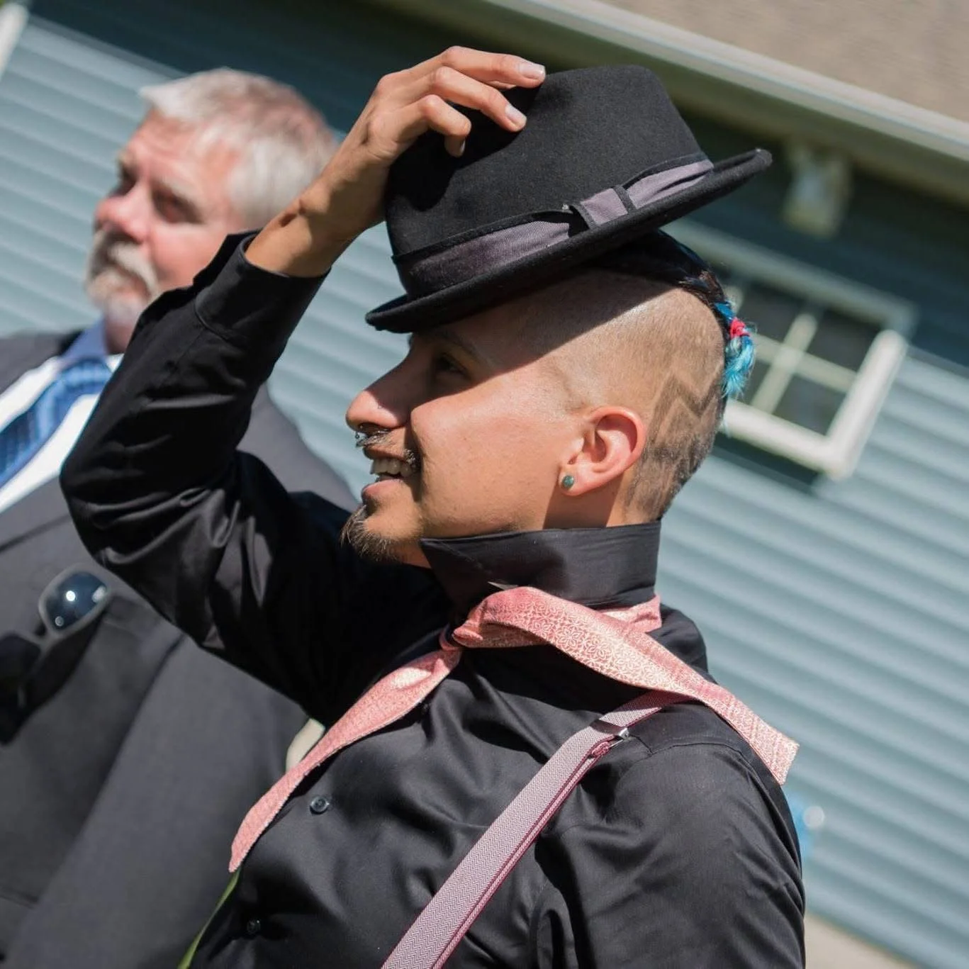 A smiling person with a mohawk hairstyle wearing a black shirt and pink suspenders, holding a black hat on their head, standing outdoors with a man in a police uniform in the background.