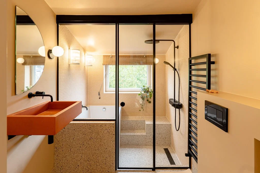 Modern bathroom with a walk-in shower, bathtub, and black-framed glass door. Features include a pink sink, round mirror, wall-mounted light, window with a plant, black towel radiator, and a cream-colored wall.