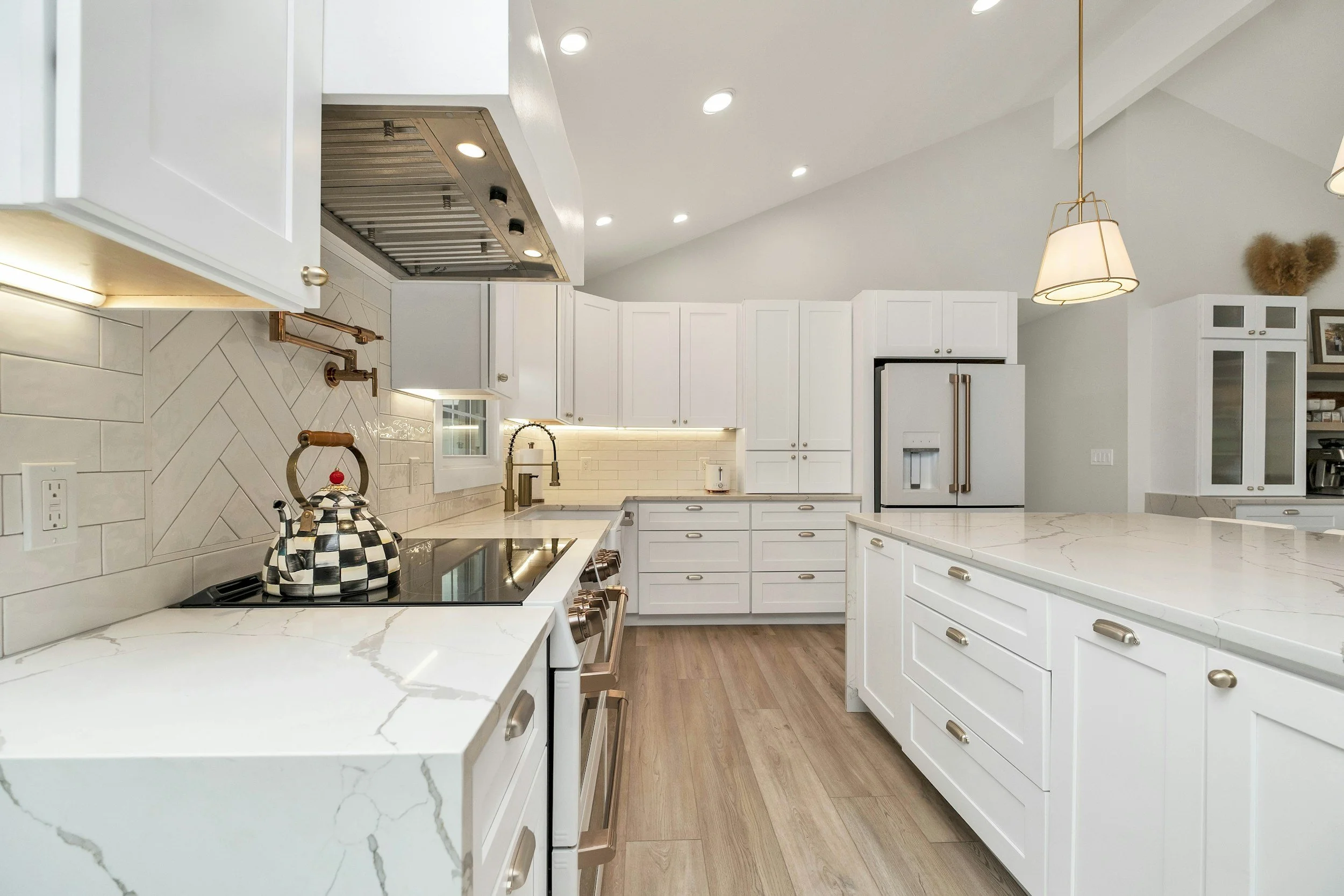 Modern white kitchen with marble countertops, wooden floors, brass hardware, and a black-and-white checkered teapot on the stove.